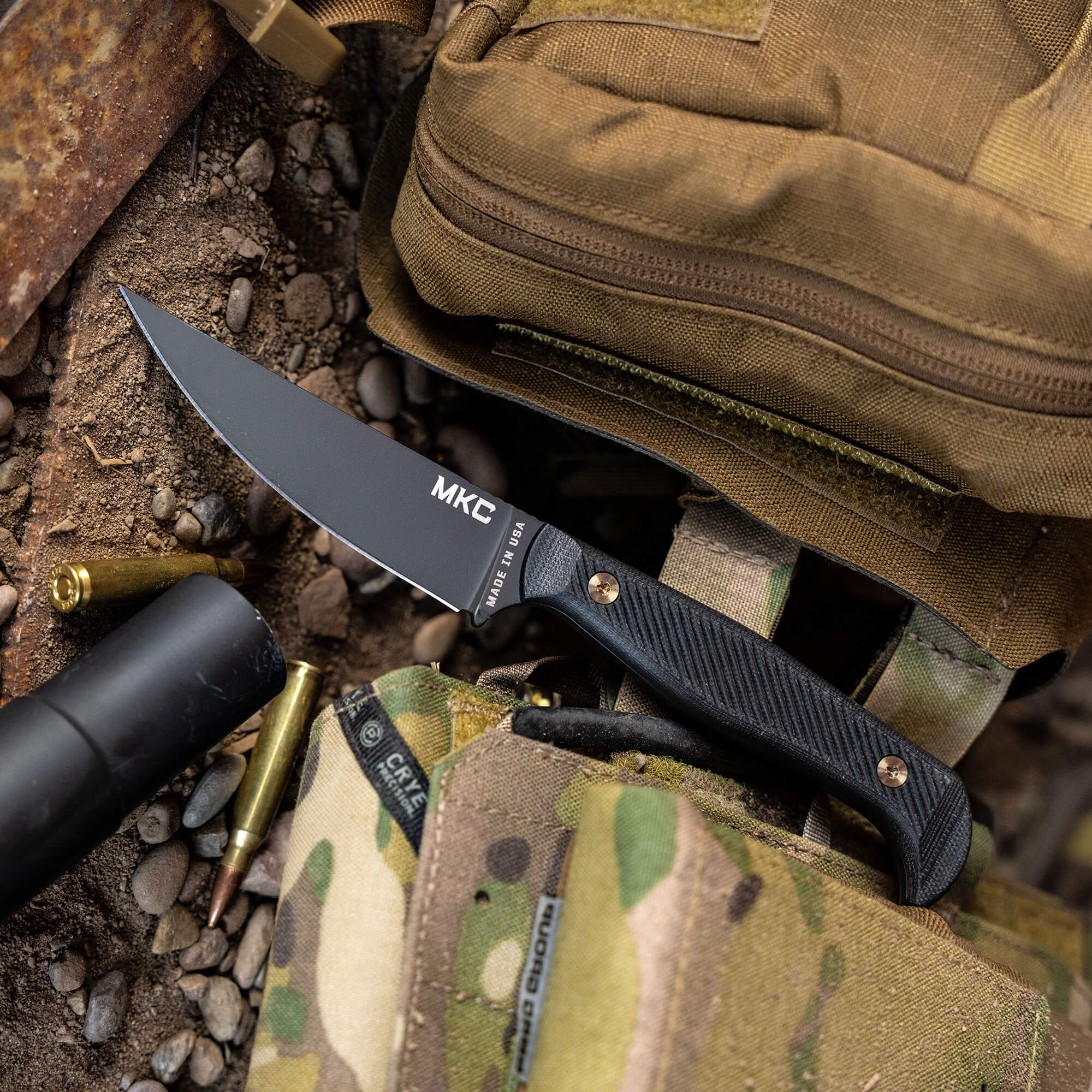 Black MKC Battle Goat tactical knife with textured handle, laid on a camouflaged military backpack among bullets and outdoor gear.