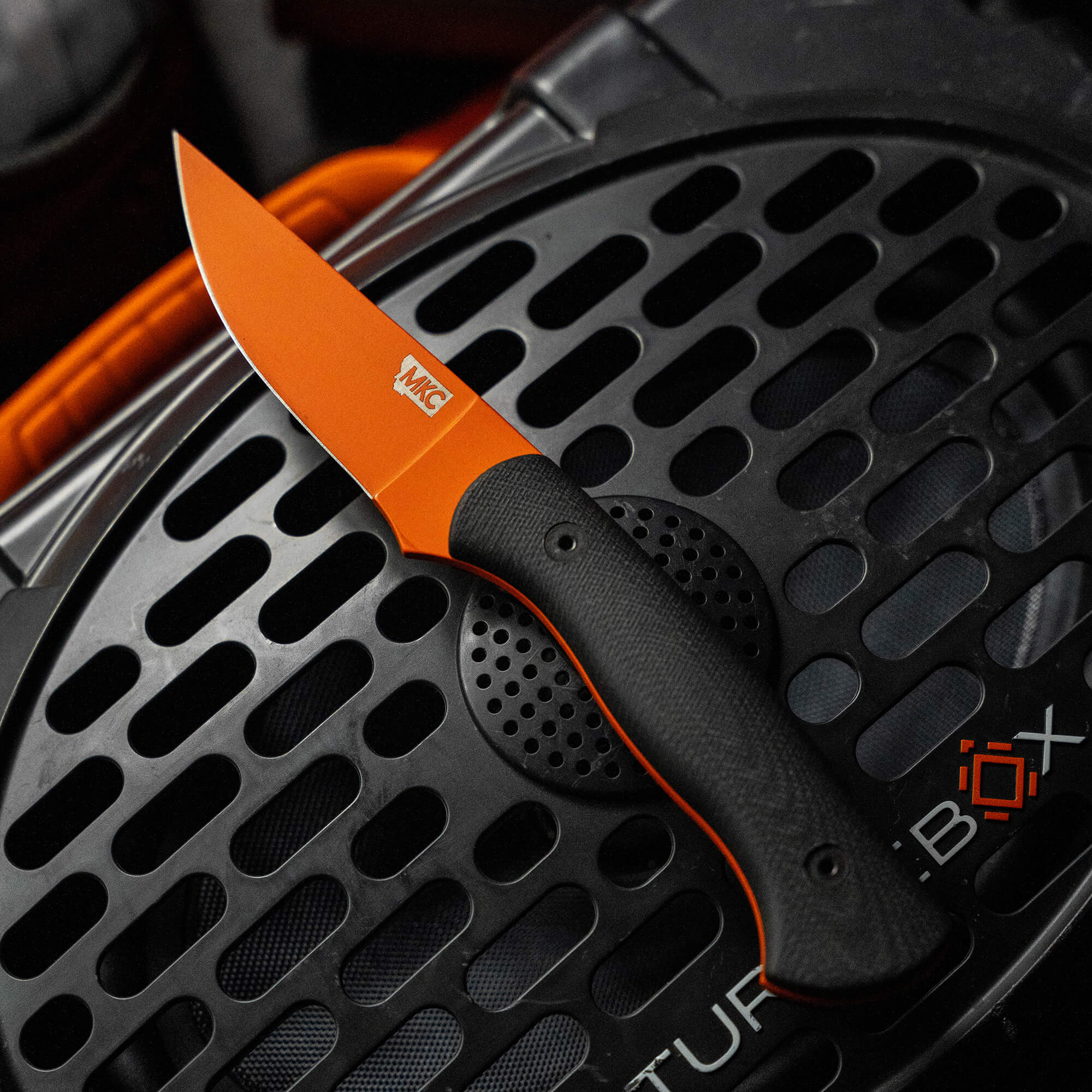 Alt text: "Blackfoot 2.0 knife with blaze orange Cerakote™ blade and black textured handle, prominently displayed on a round, patterned metal grate."