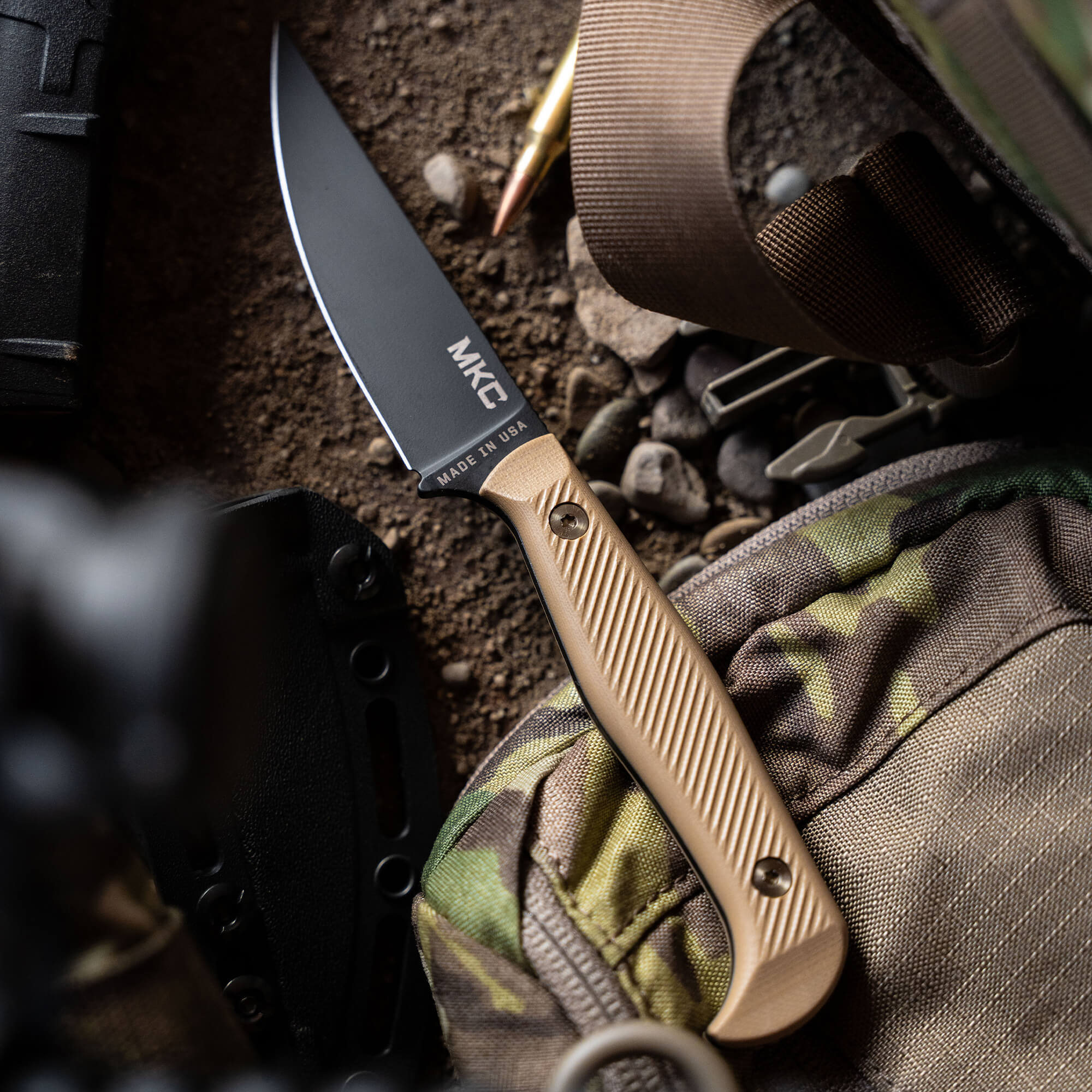 Close-up image of a tactical knife with a black blade and a coyote brown handle, labeled "MKE MADE IN USA", placed on a rocky surface with camouflage gear and tactical equipment in the background.