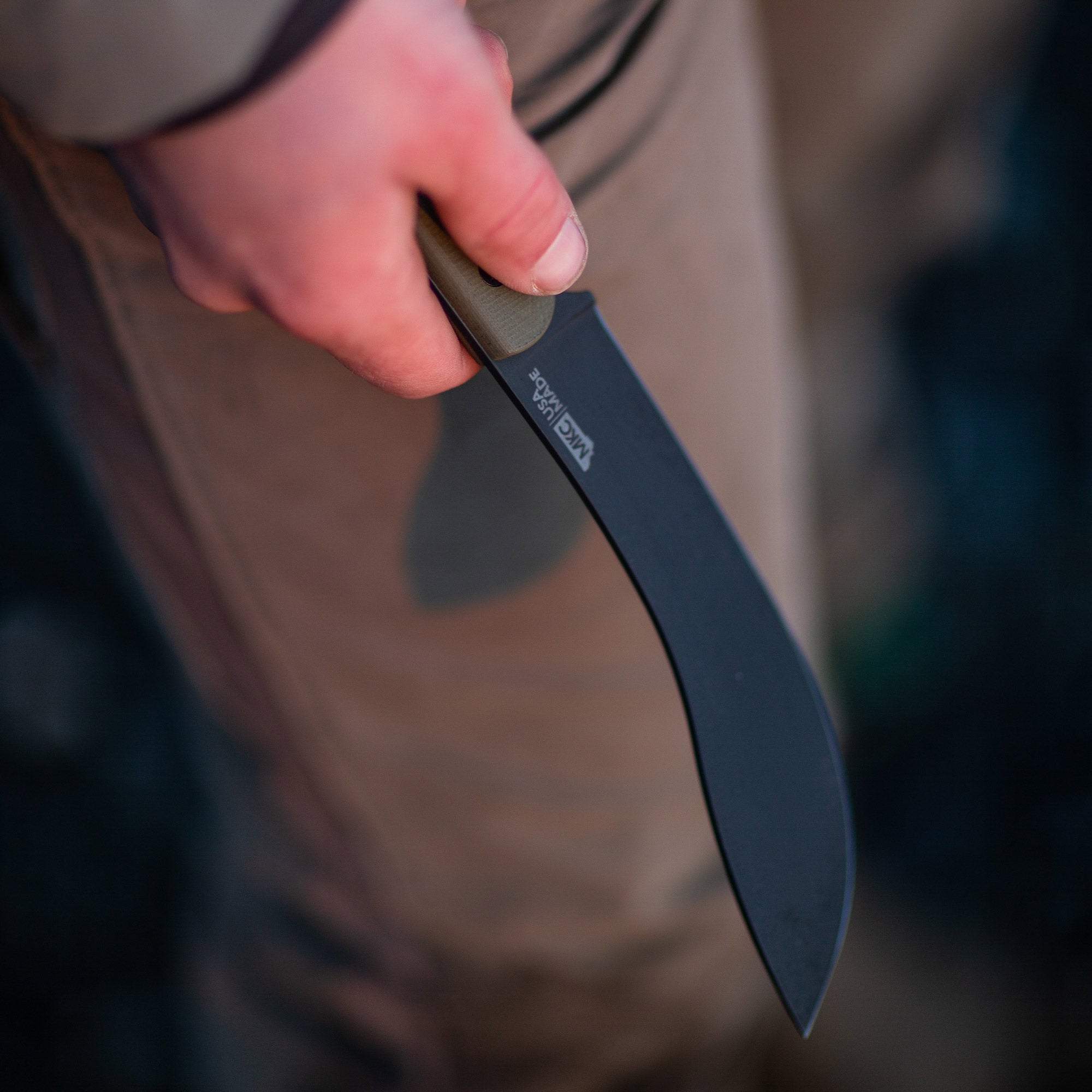 Close-up of a hand holding a Beartooth Pro Skinning Blade with an olive handle, featuring a curved black blade, against a blurred outdoor background.