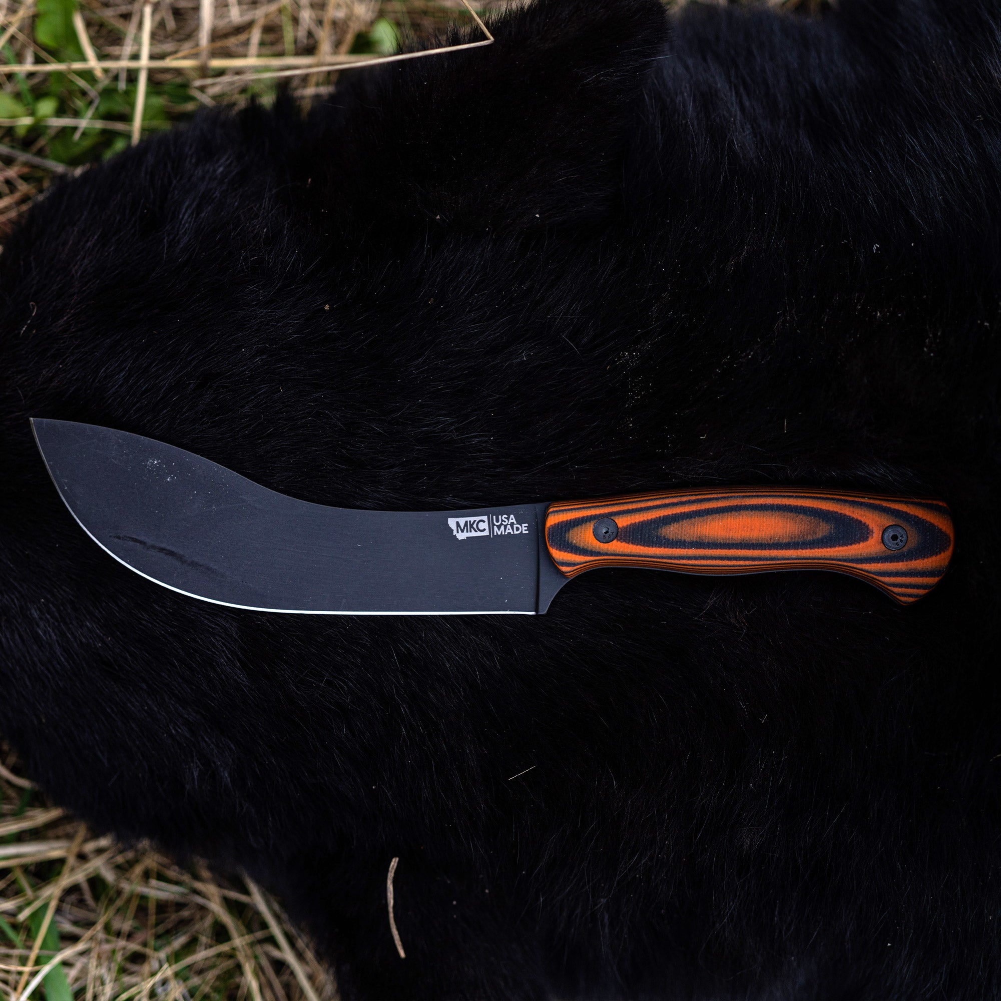 Alt text: BEARTOOTH Pro Skinning Blade with a curved black blade and an orange and black handle, displayed on a dark fur surface, emphasizing its use in hunting and skinning. Markings indicate it is made by MKC in the