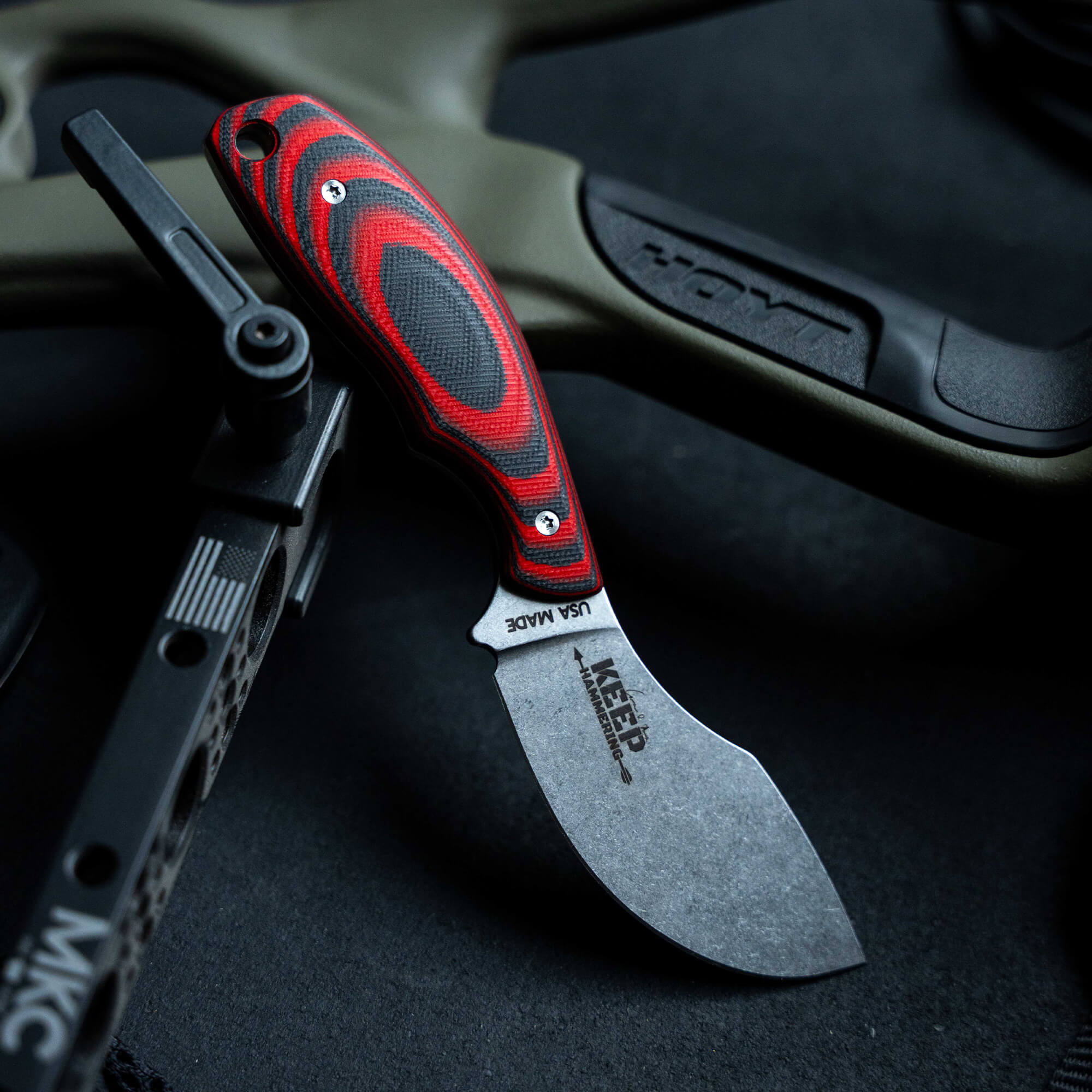 Red and black handled skinner knife with textured grip displayed on a dark background beside a compound bow, emphasizing its rugged design and utility.
