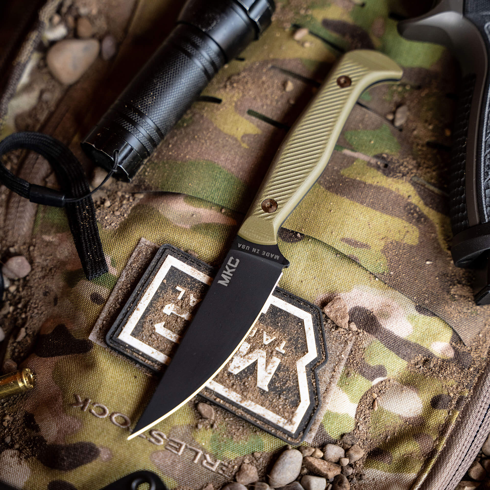 Alt text: Close-up view of the BATTLE GOAT tactical knife in black and olive drab, displayed on a camouflage background with outdoor gear elements including a flashlight and a strap.