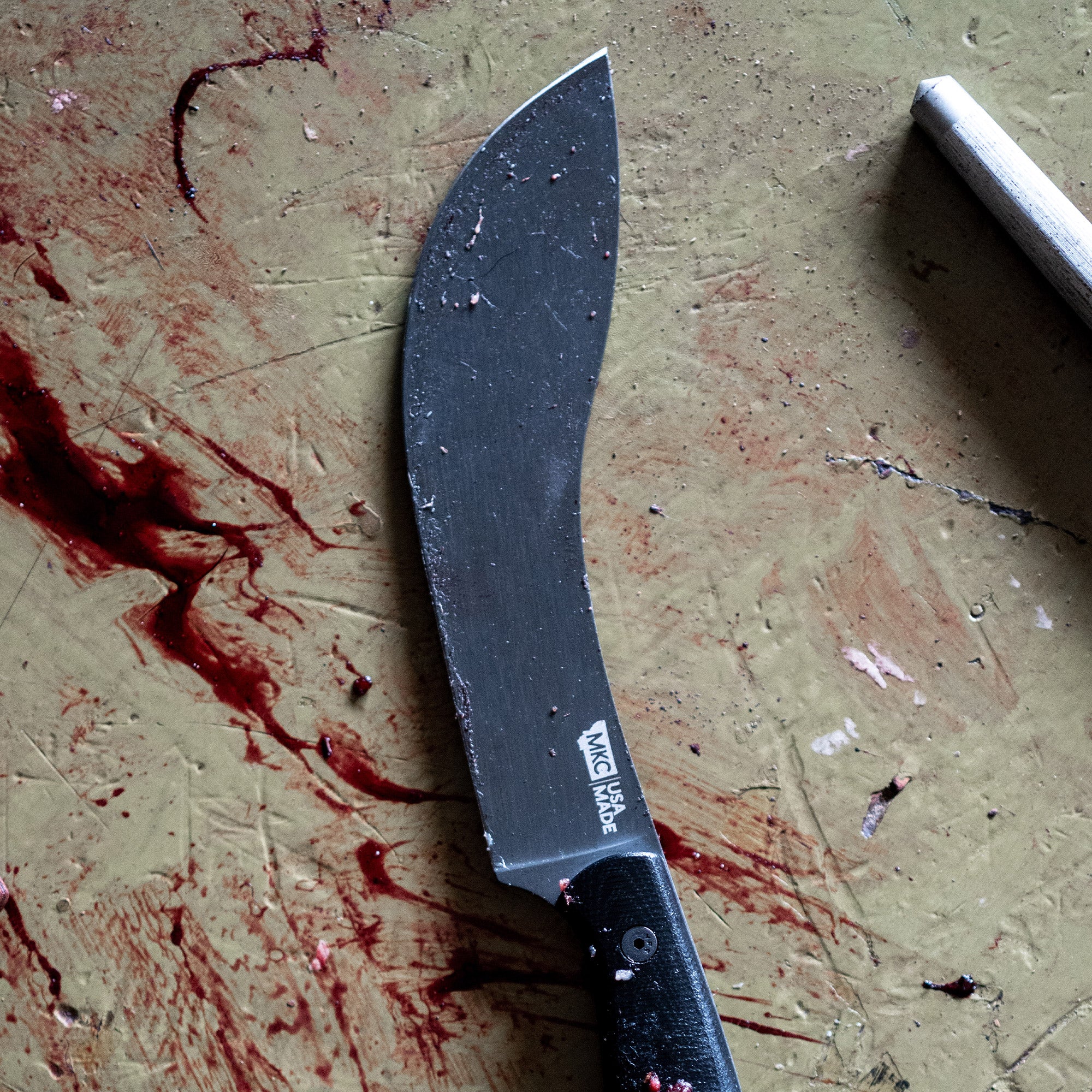 Alt text: Black Beartooth Pro Skinning Blade with a curved tip, prominently displayed on a stained and textured workshop surface, showing signs of use with visible residues and markings on the blade.