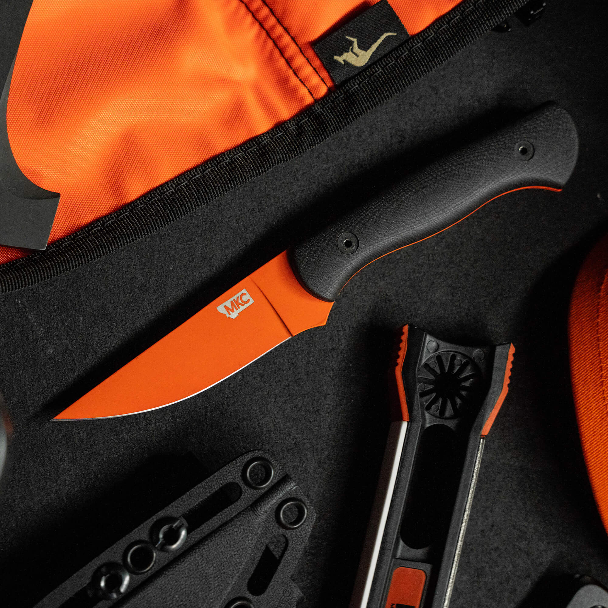 Alt text: "BLACKFOOT 2.0 knife in Blaze Orange Cerakote™ finish displayed on a black and orange surface with a hatchet, multi-tool, and sheath, illustrating outdoor gear versatility."