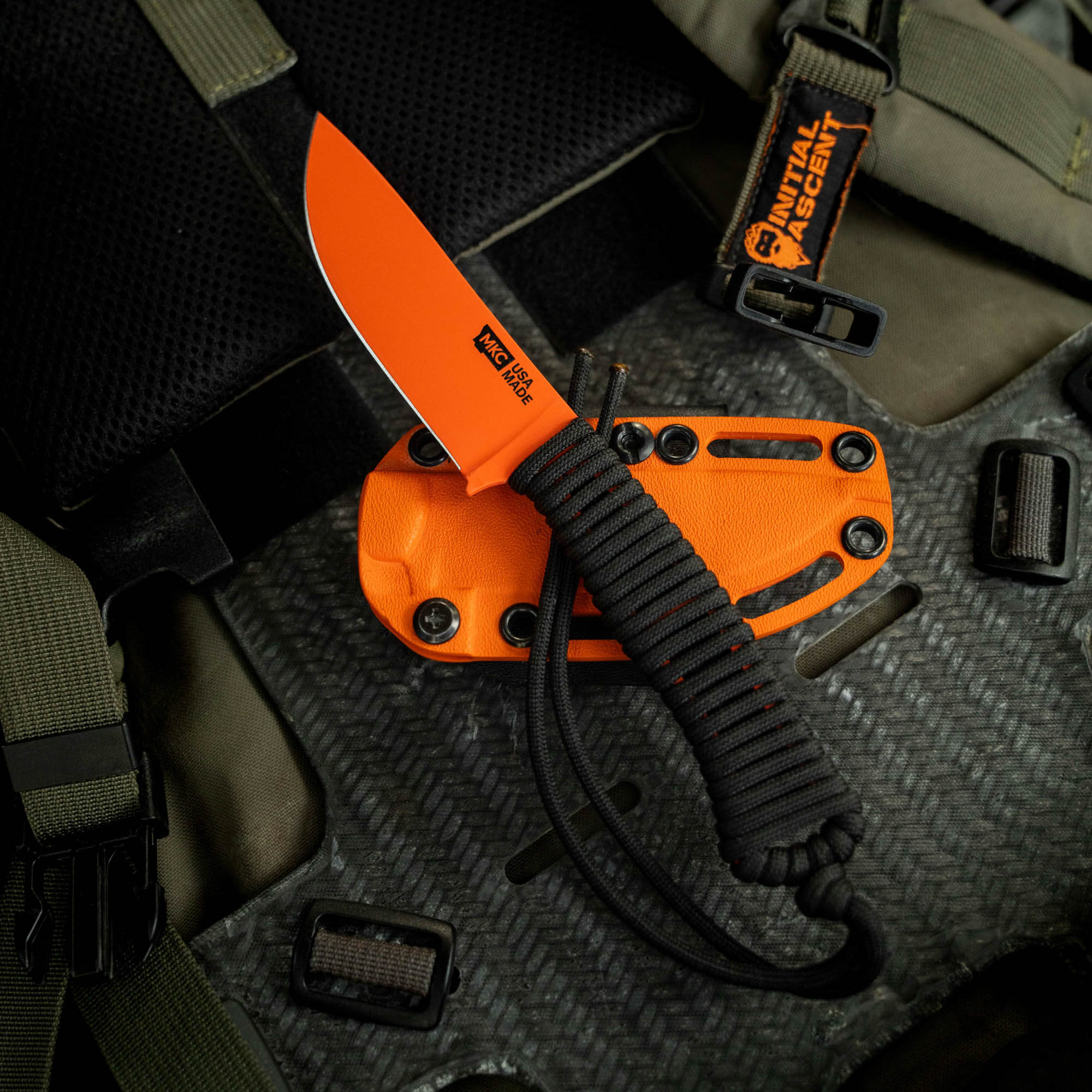Blaze orange Cerakote knife with black paracord handle, displayed in a tactical setting with a sheath.