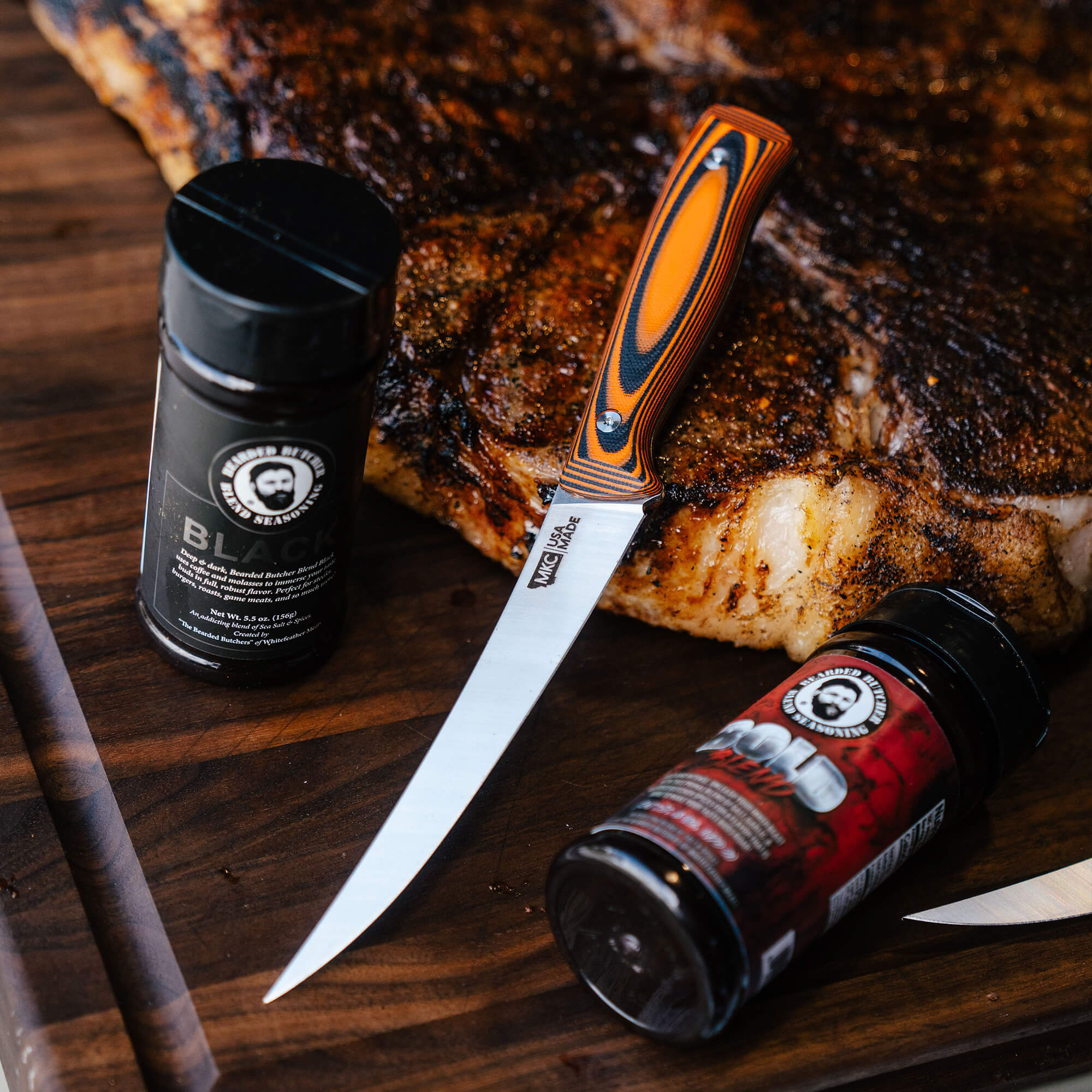 Alt text: "Bearded Butchers boning butcher knife with an orange and black handle on a wooden surface next to two spice containers, with a cooked steak in the background."