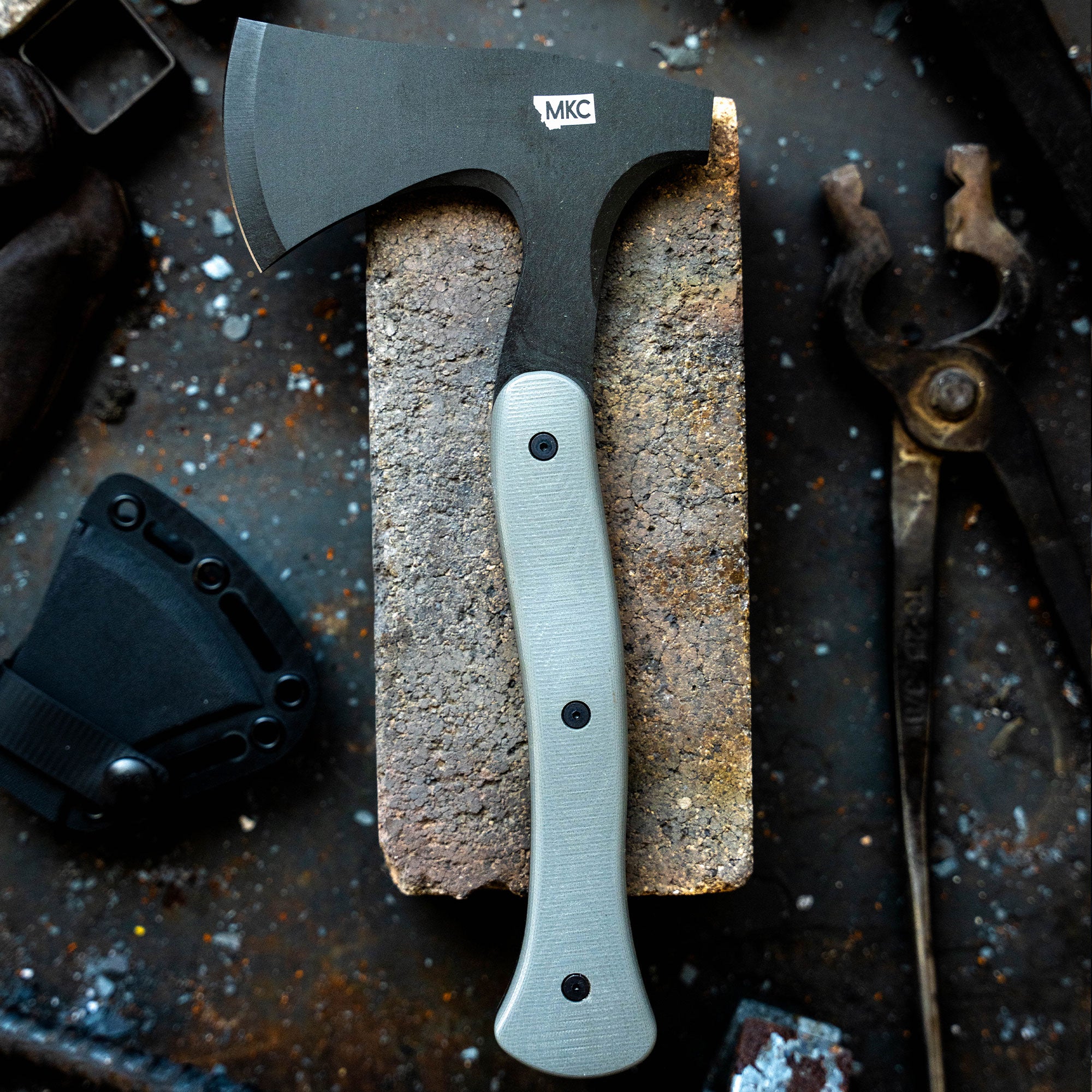 Alt text: Closeup of AEV Hellgate Hatchet from MKC, showcasing its robust black steel blade and grey handle, displayed on a textured stone surface with rustic tools in the background.
