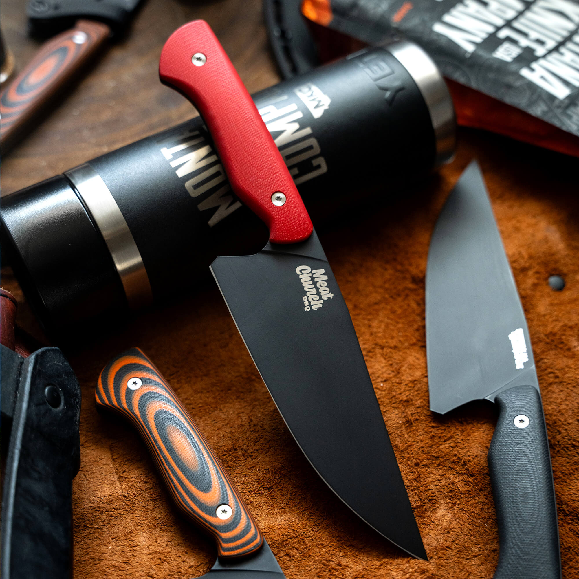 Alt text: "Close-up view of a Meat Church chef knife with a red handle and black blade, prominently displayed next to a cylindrical container and other knives on a textured brown surface, highlighting the exclusive and sold out 'Secret Drop' product."