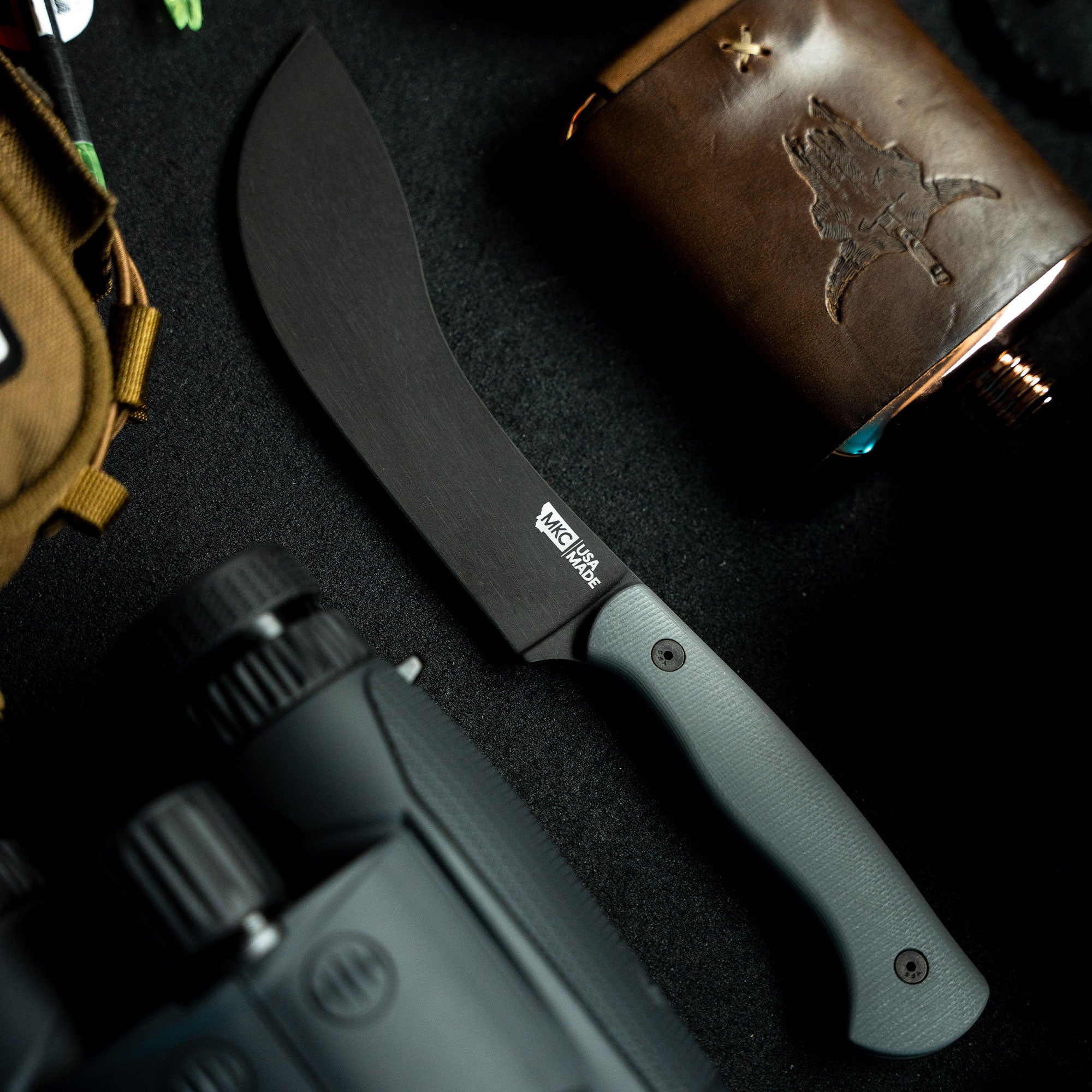 Alt text: "BEARTOOTH Pro Skinning Blade in grey, featuring a curved black blade and a textured grey handle, displayed on a dark background with hunting accessories including a leather pouch and binoculars."