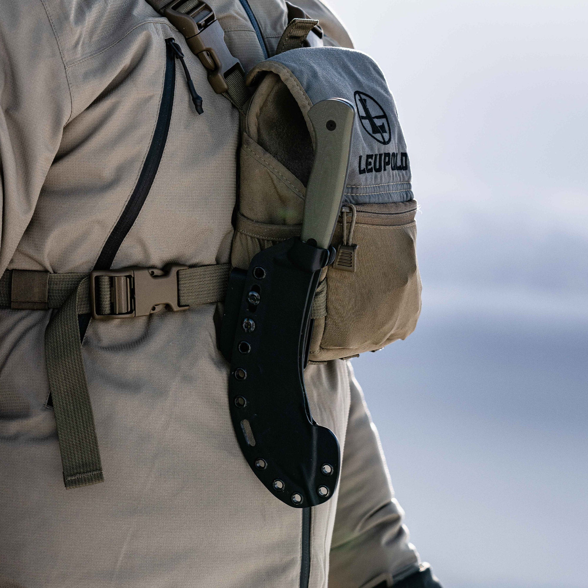 Alt text: "The Beartooth Pro Skinning Blade in olive color attached to the waist strap of a beige tactical vest, showing the blade in its black sheath."