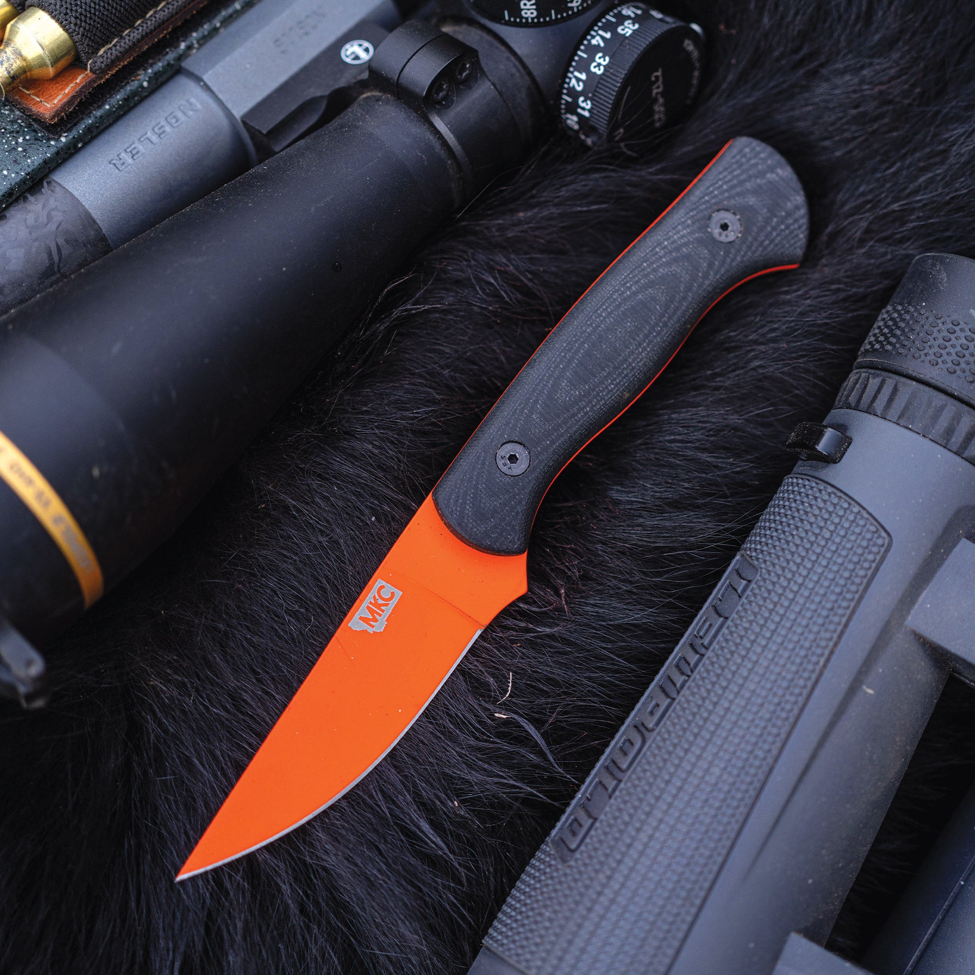 Alt text: "Close-up of a Blackfoot 2.0 hunting knife with a blaze orange Cerakote blade and black handle, lying on a black fur surface next to a rifle and a scope."