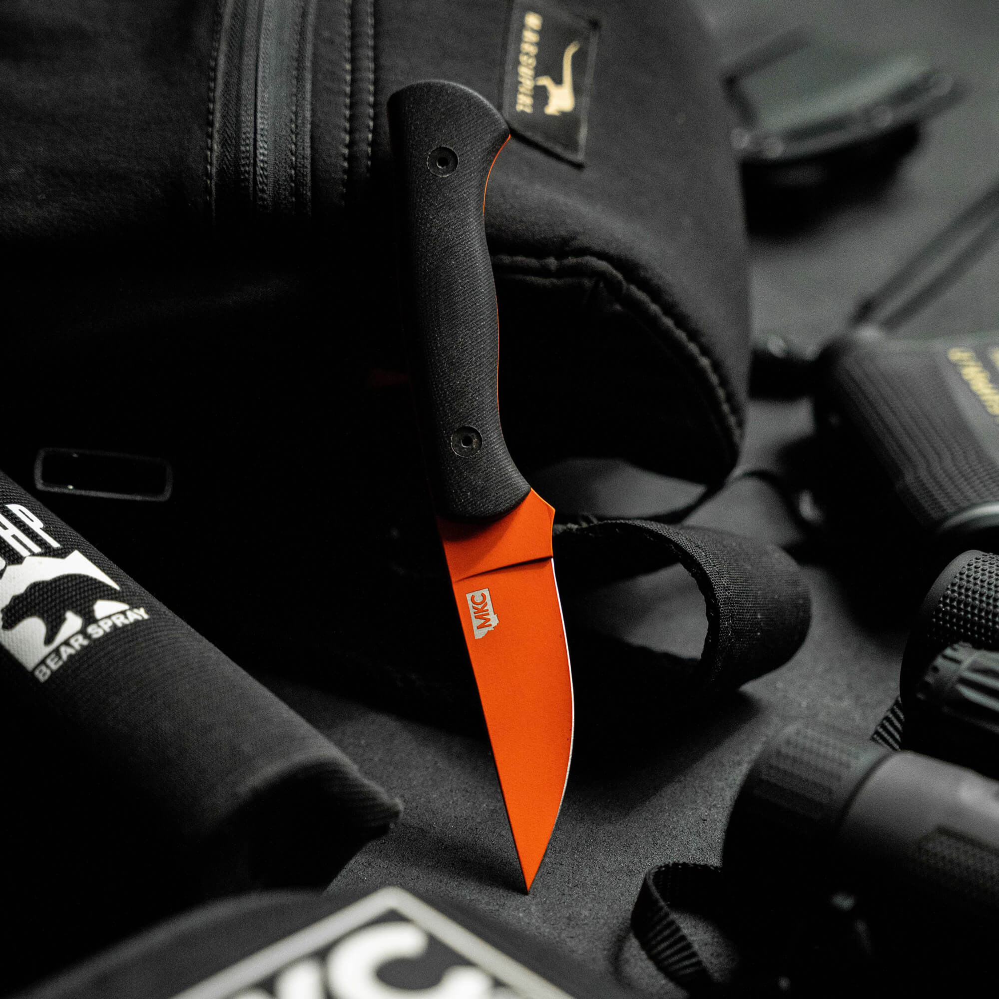 Alt text: "BLACKFOOT 2.0 tactical knife with a blaze orange Cerakote blade and black handle, displayed on a tactical gear background including a tactical vest and other equipment."