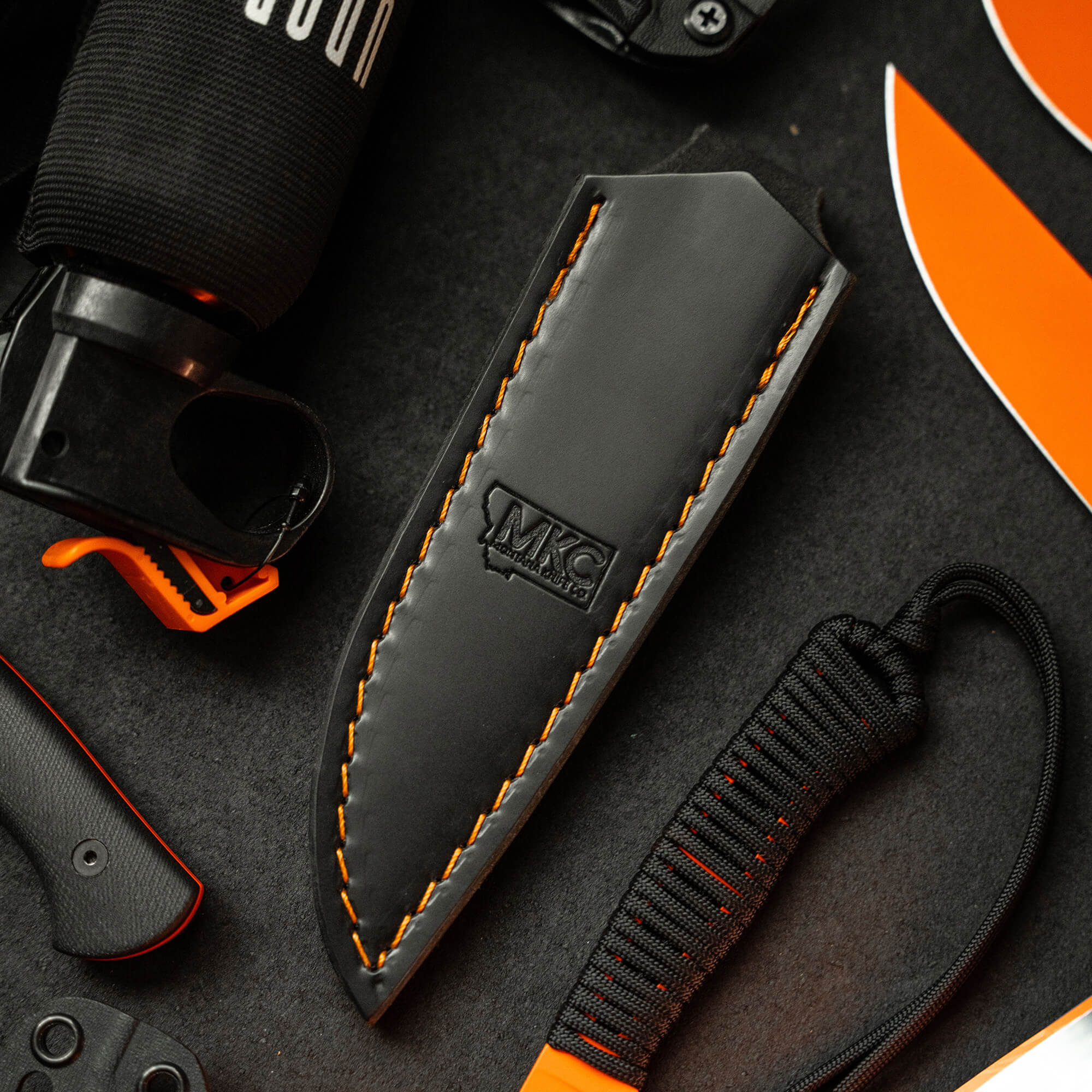 Black leather sheath for vertical belt carry with orange stitching and MKC logo, displayed amongst various outdoor gear including a flashlight and knife.