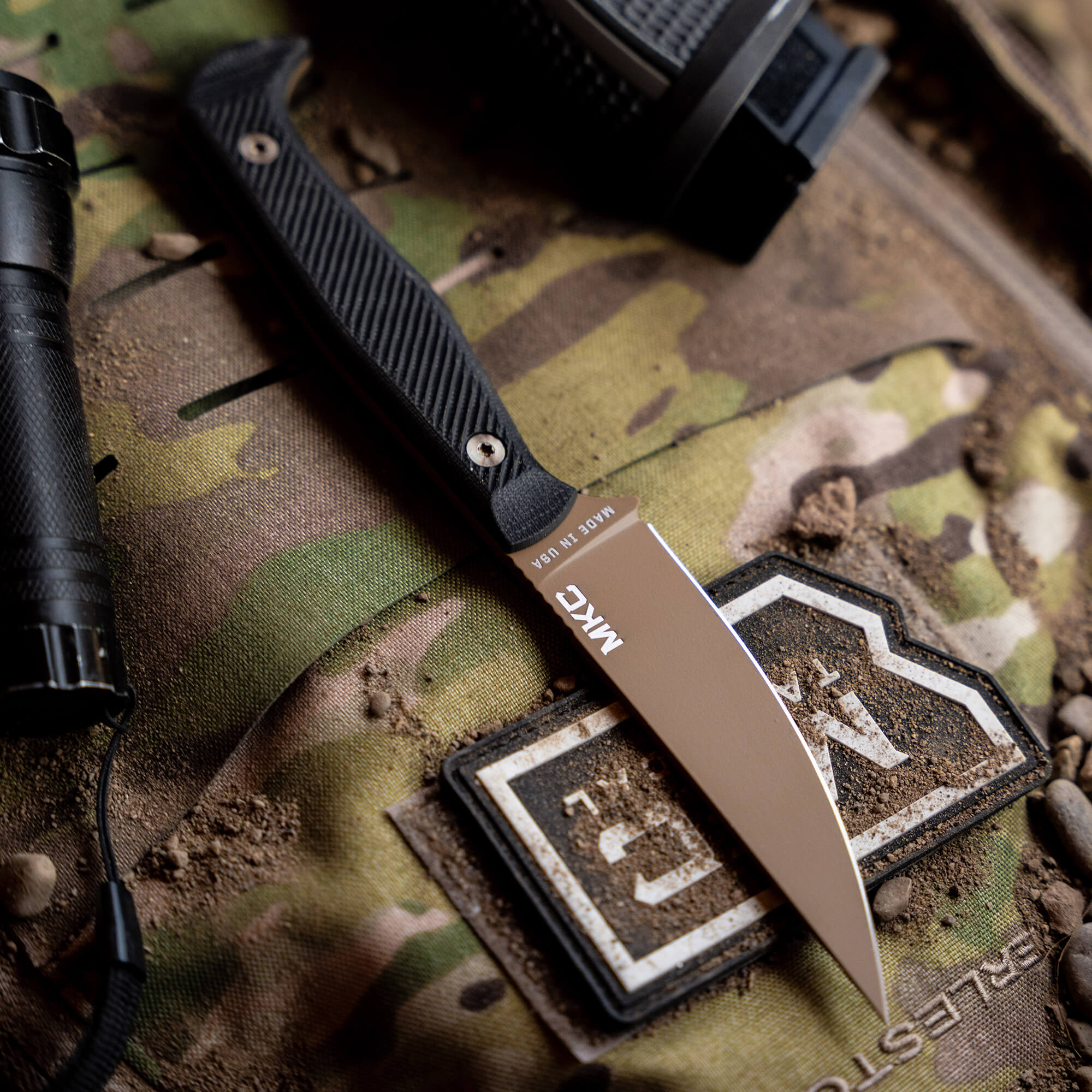 Alt text: "Close-up view of a BATTLE GOAT tactical knife in coyote black color with a focused shot on the blade, displayed on a camouflage background alongside outdoor gear including a flashlight."