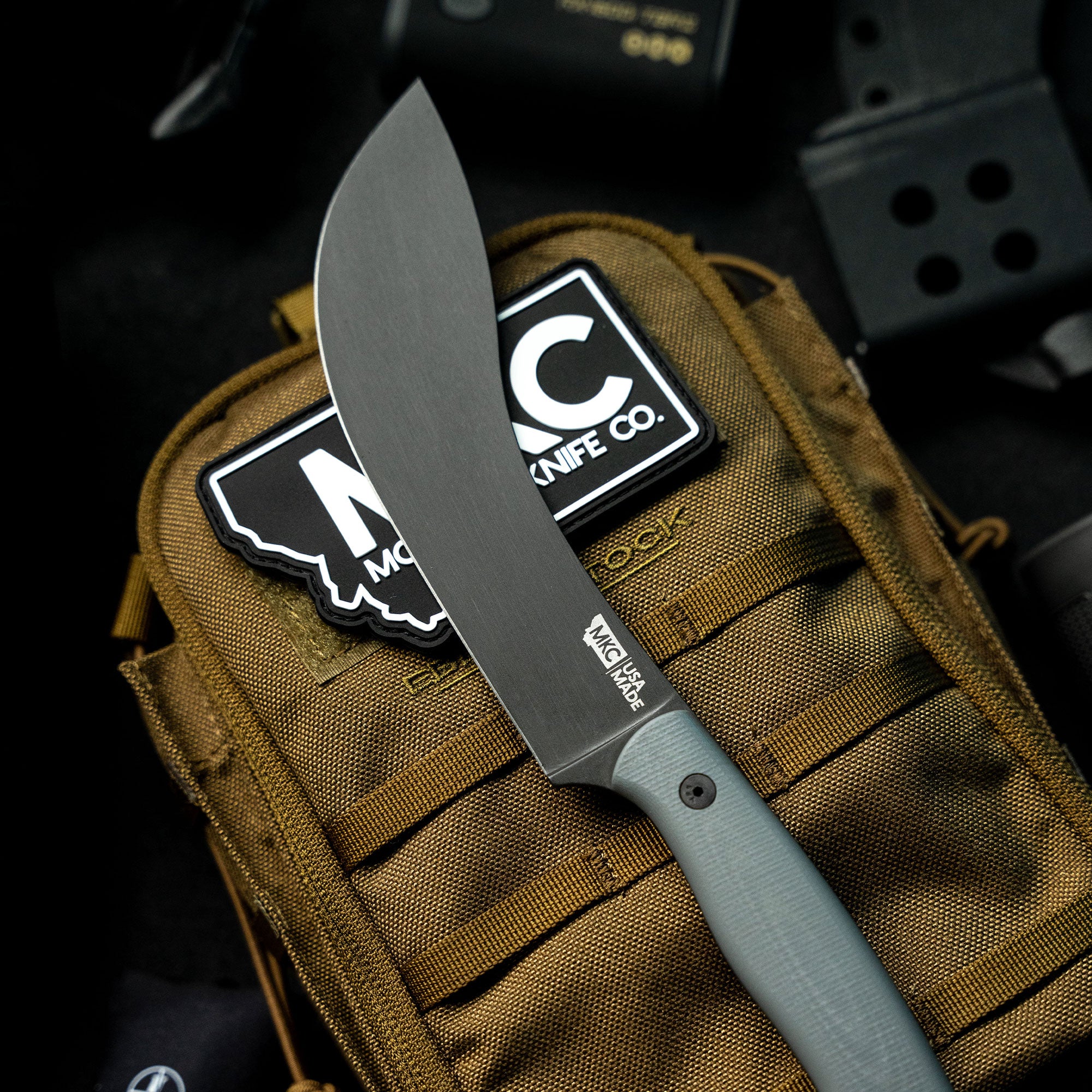 Alt text: "Close-up of the Beartooth Pro Skinning Blade in grey, with a thick, sturdy blade displayed against a brown tactical backpack adorned with patches from the Montana Knife Company."