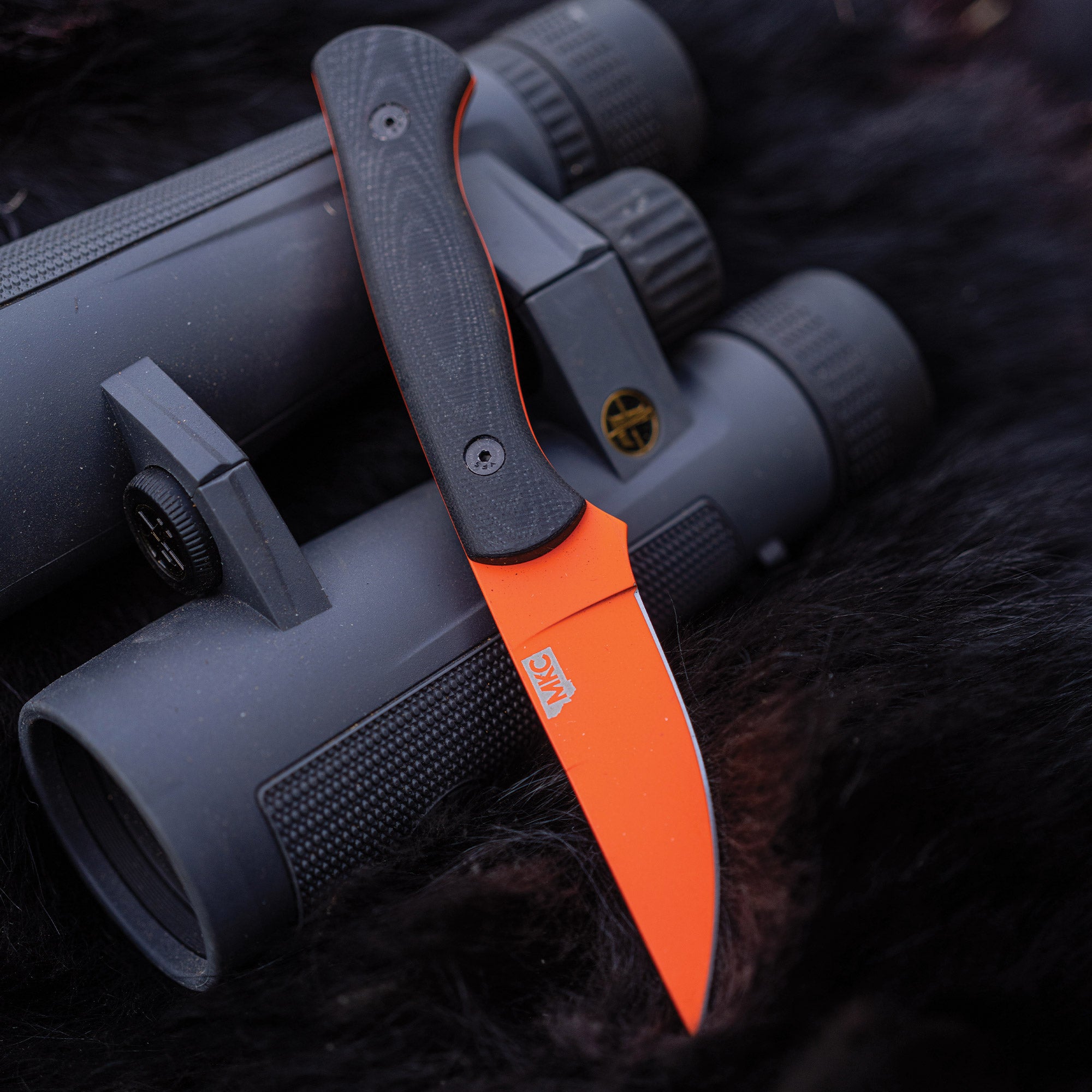 Blaze orange fixed blade knife with black handle partially resting over the top of two gray binoculars, displayed on a fur surface.