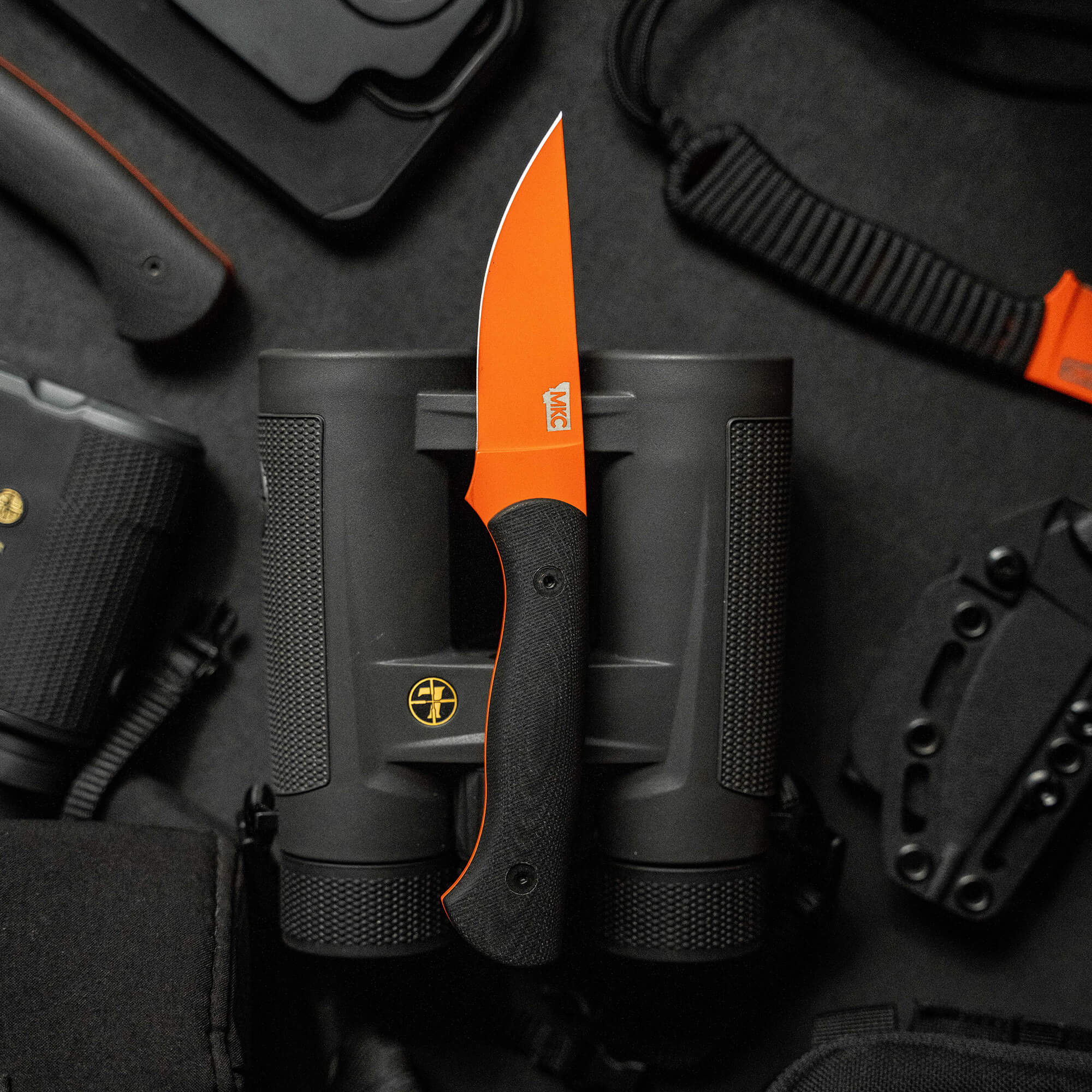 Alt text: "BLACKFOOT 2.0 knife with a vibrant blaze orange Cerakote blade, nestled in a black secure sheath, surrounded by various tactical gear including holsters and binoculars on a dark background."