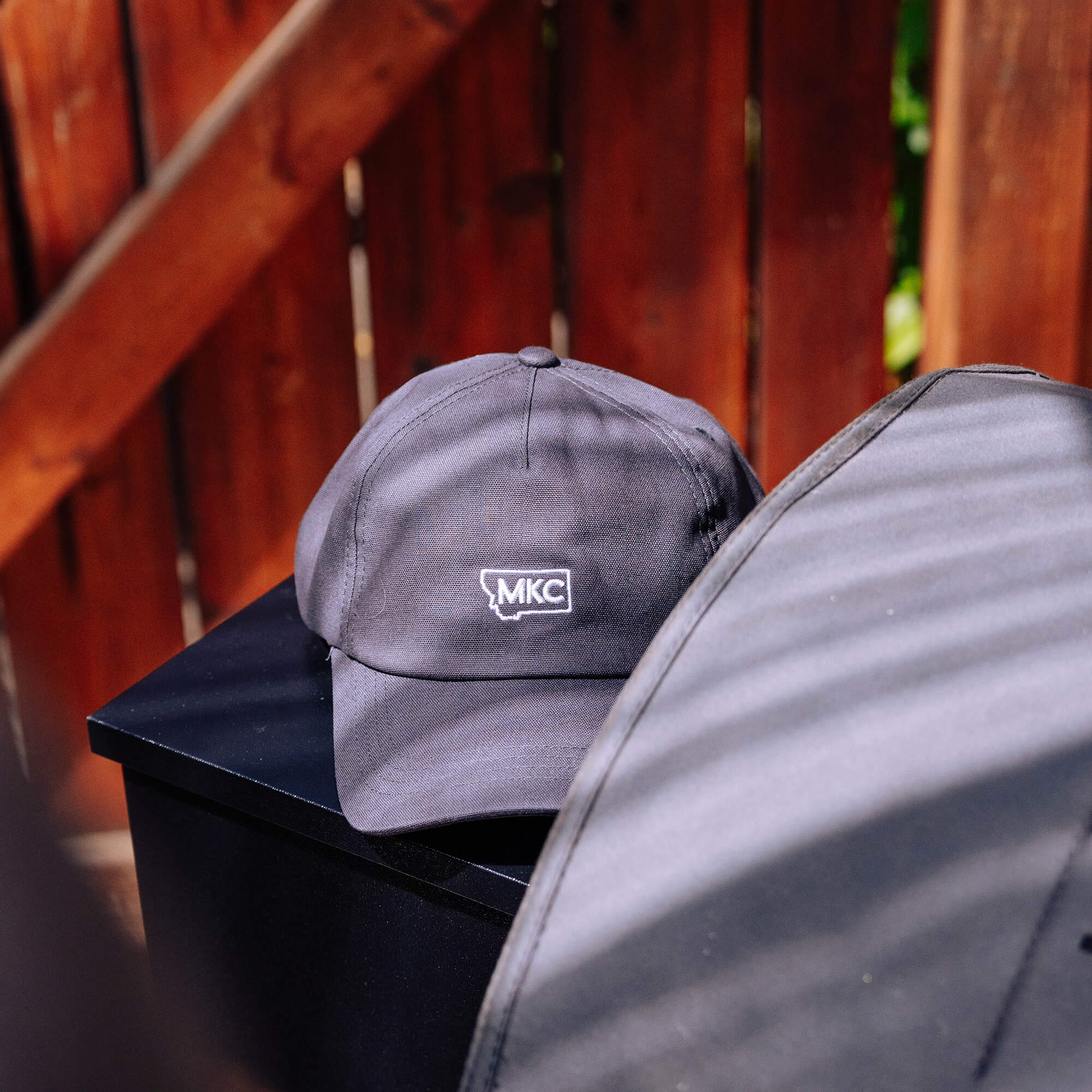 Grey MKC canvas dad hat featuring a logo, placed on a black surface with a wooden backdrop. Ideal for outdoor enthusiasts.