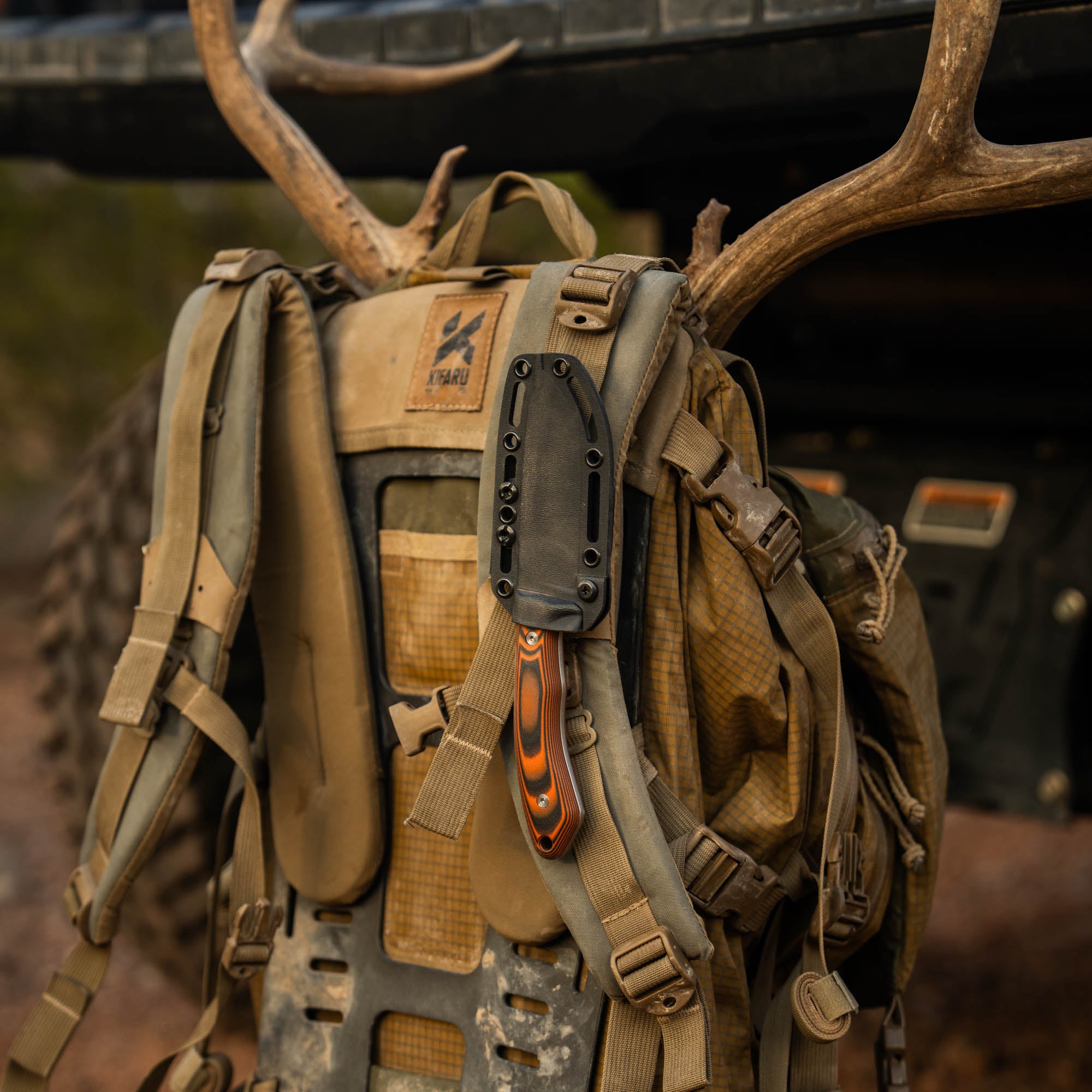 MKC Mule Deer Knife in orange and black, attached to a rugged backpack with antlers in the background, ideal for outdoor use.