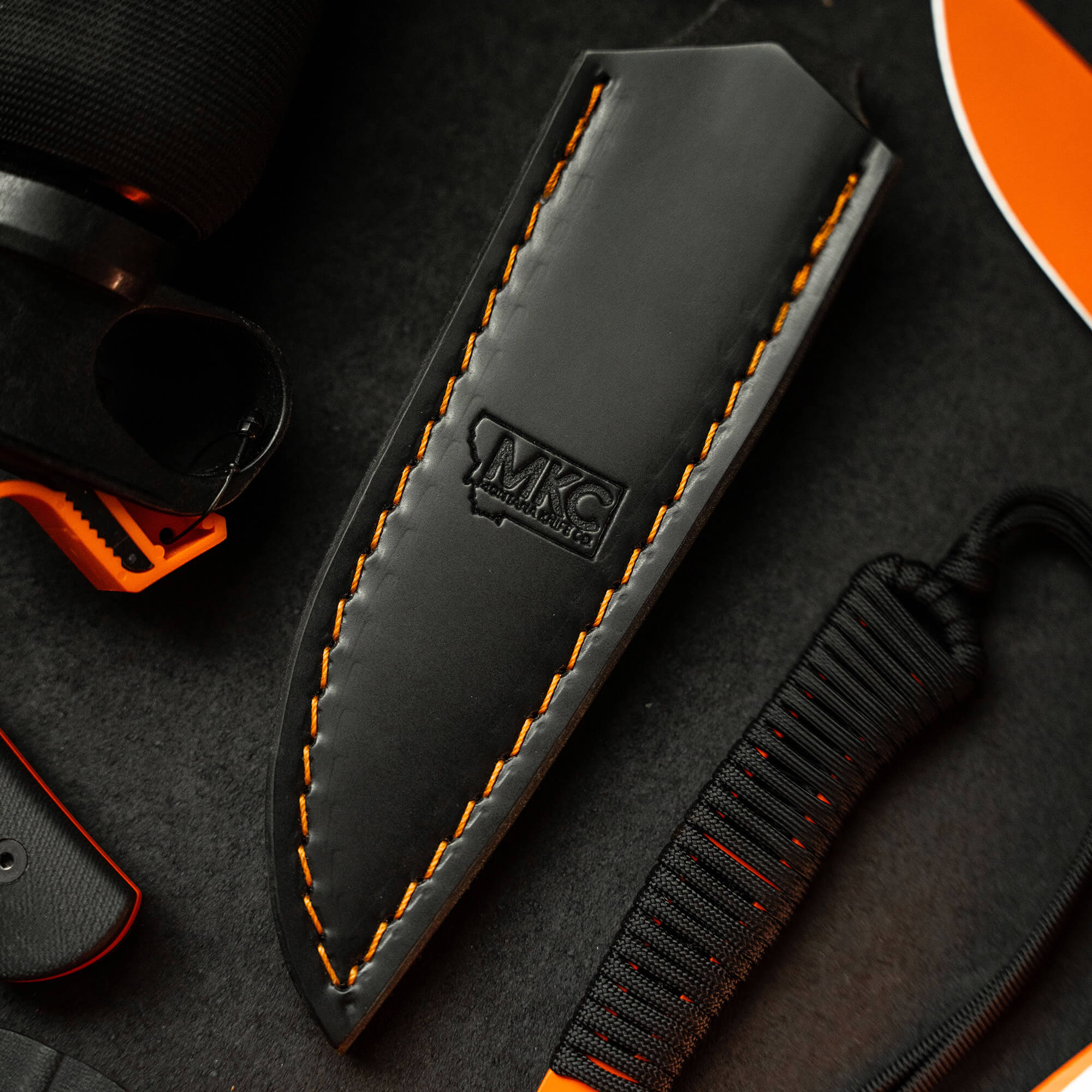 Alt text: "Black leather sheath with orange stitching for vertical belt carry, featuring the embossed MKC logo, displayed on a dark textured background alongside orange and black outdoor gear elements."