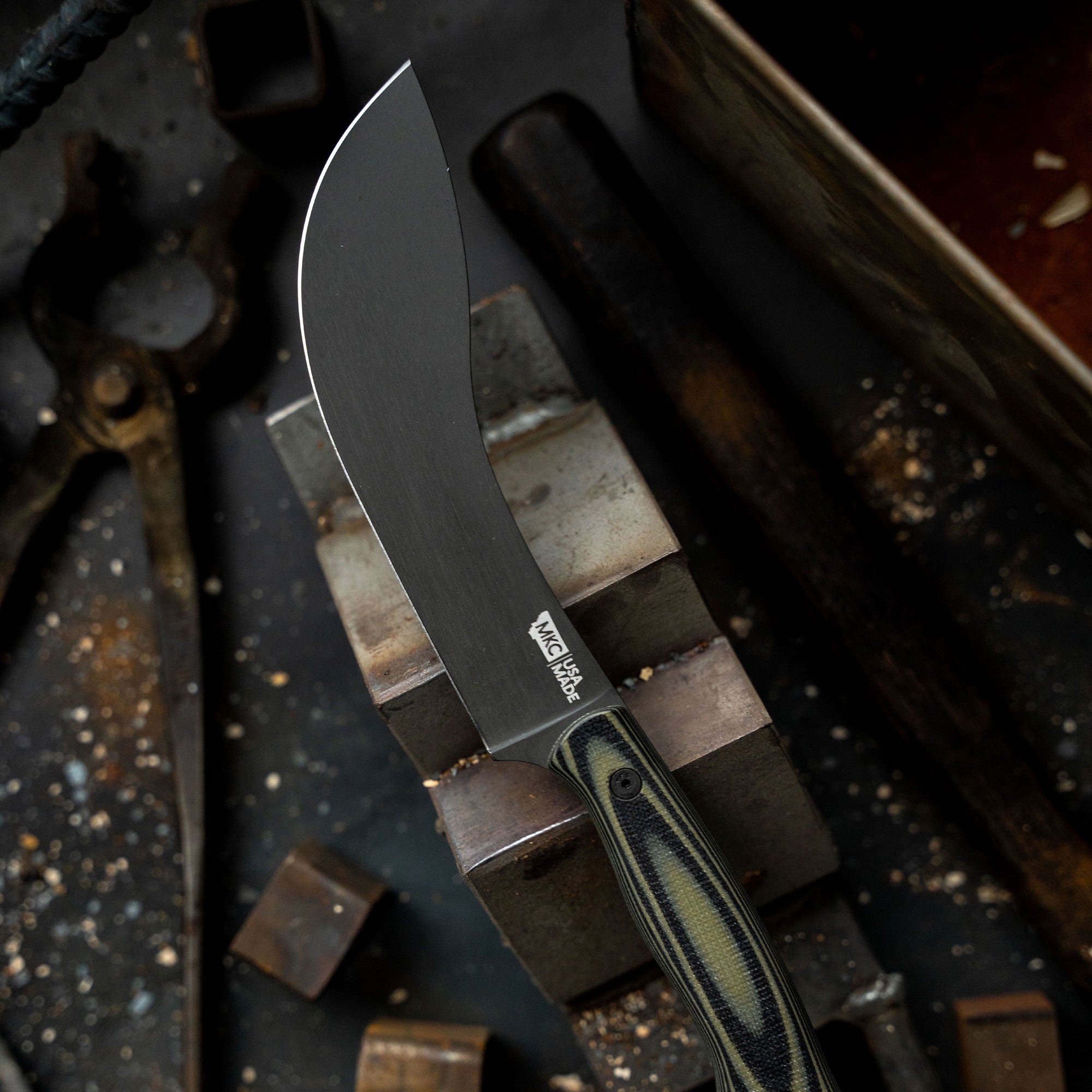 Alt text: Close-up of a Beartooth Pro Skinning Blade with a curved black blade and a striped green and black handle, displayed on a rustic workshop setting with metal tools and wooden blocks in the background.
