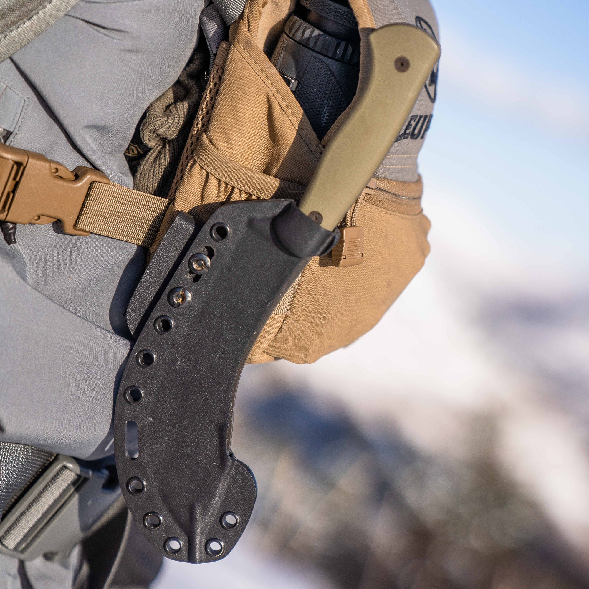 Close-up image of a Beartooth Pro Skinning Knife with an olive handle attached to a tactical backpack, featuring a black sheath, in a snowy outdoor setting.