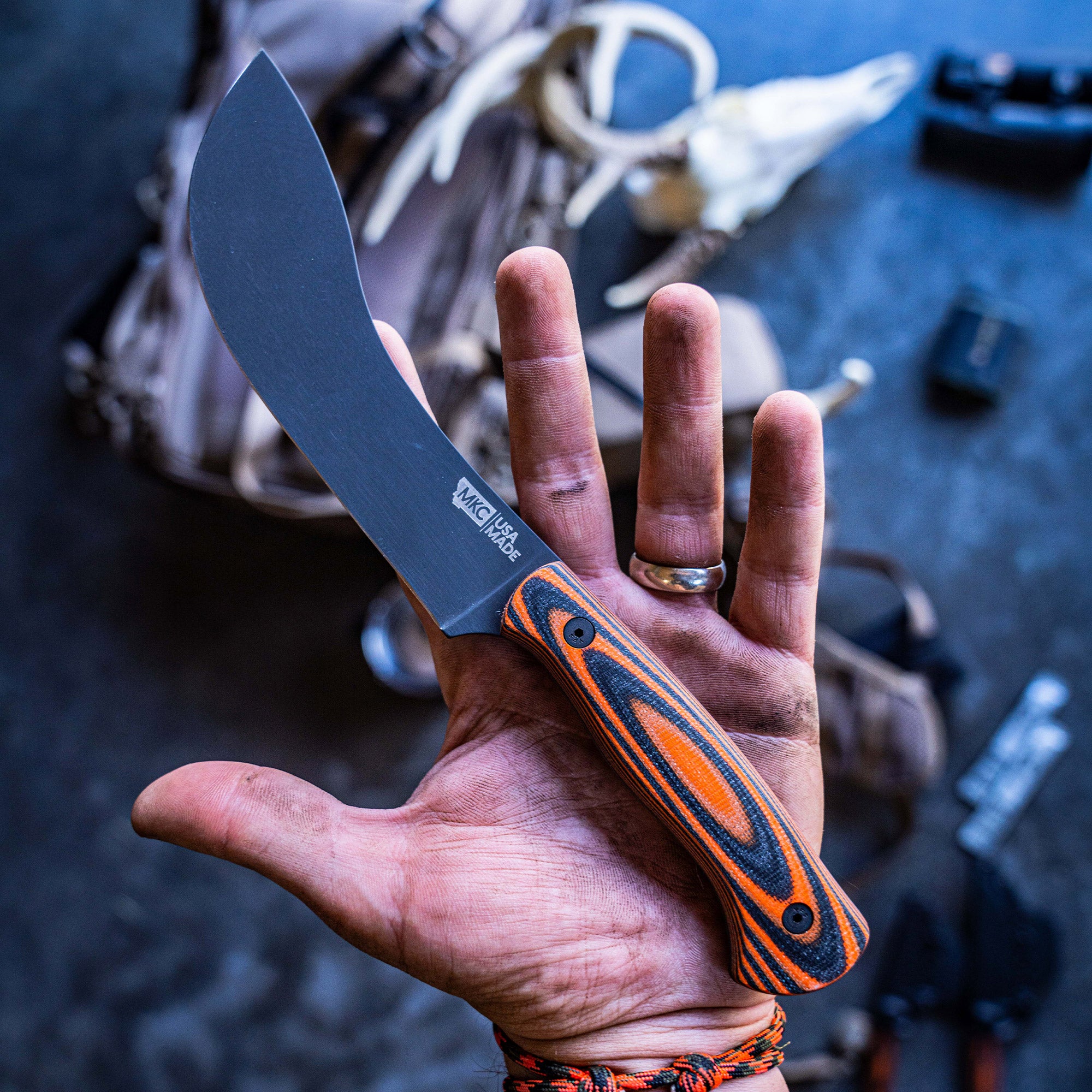 Alt text: Close-up of a Beartooth Pro Skinning Blade with orange and black handle held in a person's hand, showing the blade's matte finish and ergonomic design, set against a blurred background with outdoor gear.
