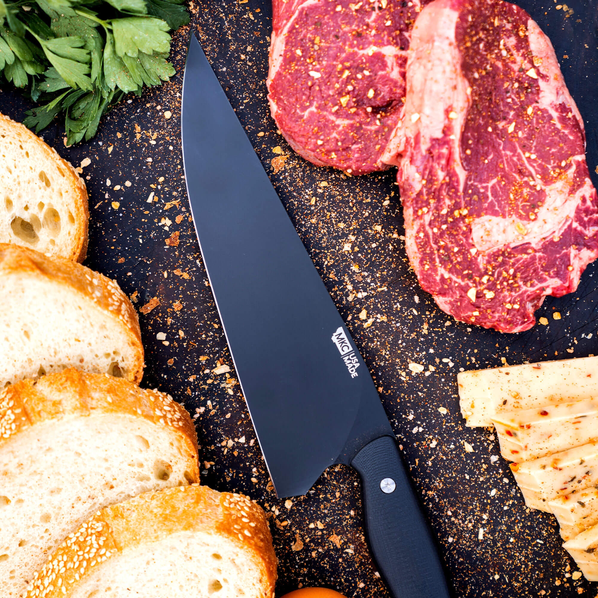 Alt text: "Professional BIGHORN CHEF knife in black, displayed on a dark countertop alongside raw marbled meat, sliced bread with sesame seeds, and herbs, highlighting the knife's sleek design and culinary versatility."