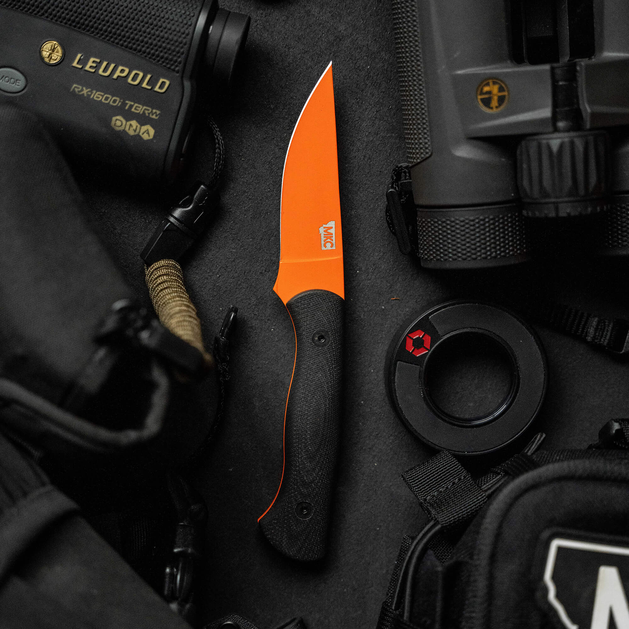 Alt text: "Blackfoot 2.0 fixed blade knife with a vibrant blaze orange Cerakote™ finish and a black handle, displayed among various tactical gear including a Leupold rangefinder and a tactical backpack."