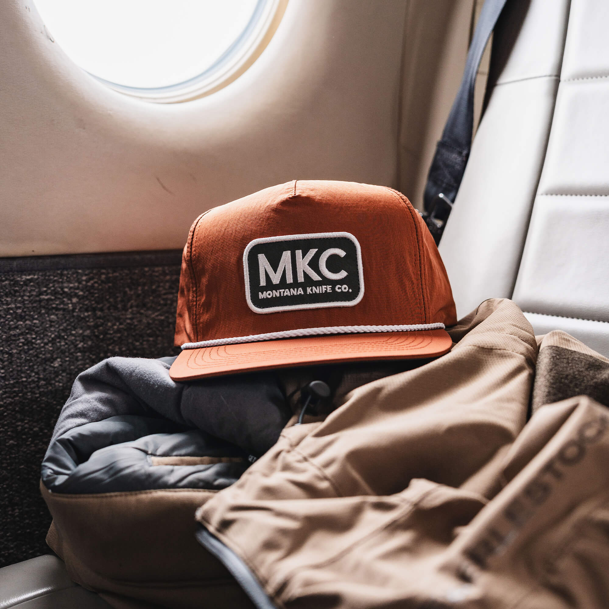 Rust-colored MKC River Rope Hat resting on a jacket inside an airplane, showcasing its outdoor-ready design.