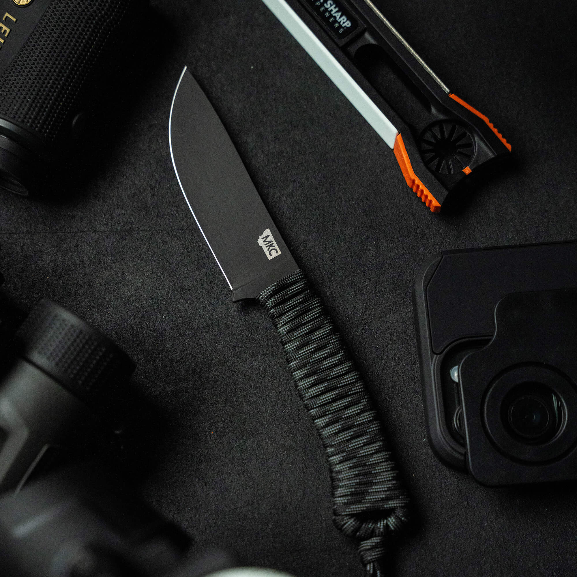 Alt text: "Close-up view of the 'Stoned Goat 2.0' knife by Wild Sheep Foundation, featuring a sturdy black blade and a paracord-wrapped handle, displayed alongside outdoor gear including a compact digital camera and precision twe