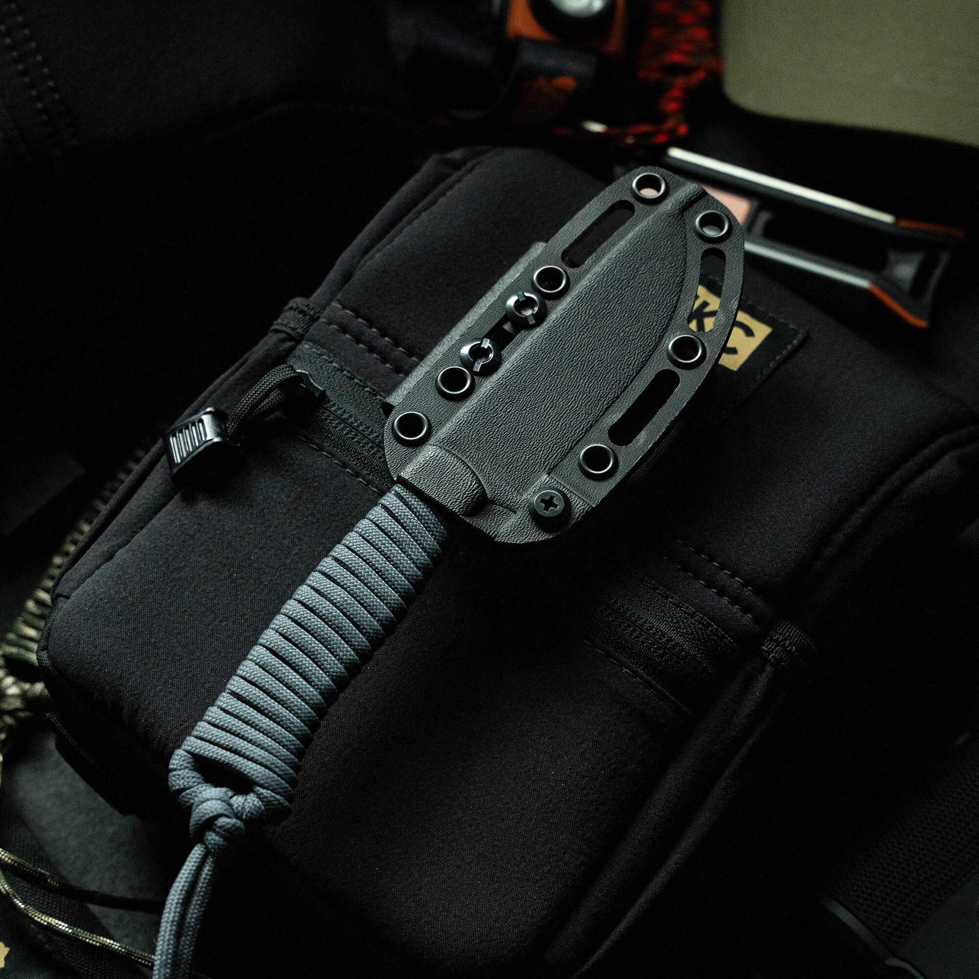 Alt text: Close-up view of a Stoned Goat 2.0 knife by Wild Sheep Foundation, featuring a sleek black handle and sheath attached to a tactical bag, with a gray paracord tied to it.