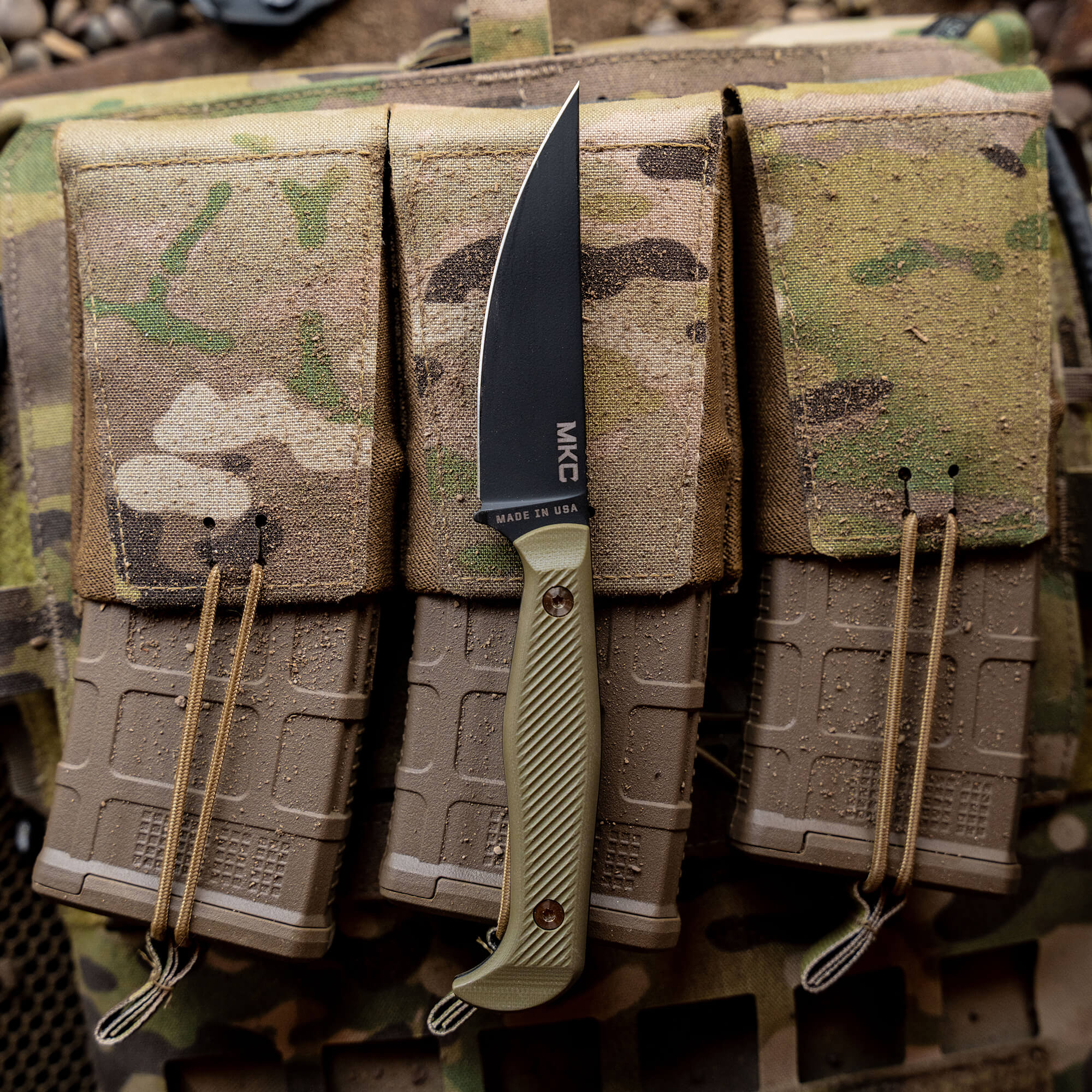 Alt text: "MKC Battle Goat tactical knife with a black blade and olive drab handle, displayed vertically against a camouflage and tan tactical gear background, highlighting its military-grade design and 'Made in USA' label."
