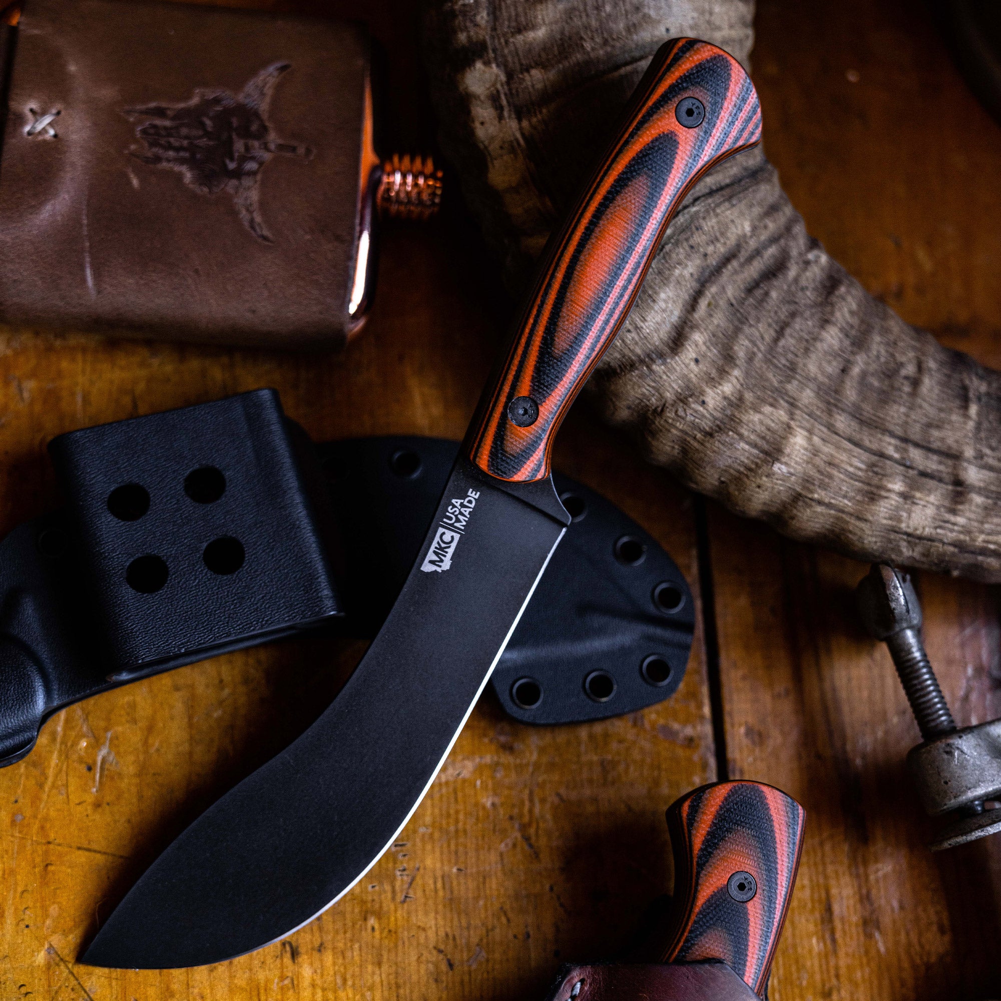Alt text: "Beartooth Pro Skinning Blade with a black coated blade and a striped orange and black handle, displayed alongside its black sheath on a rustic wooden surface, surrounded by vintage accessories."