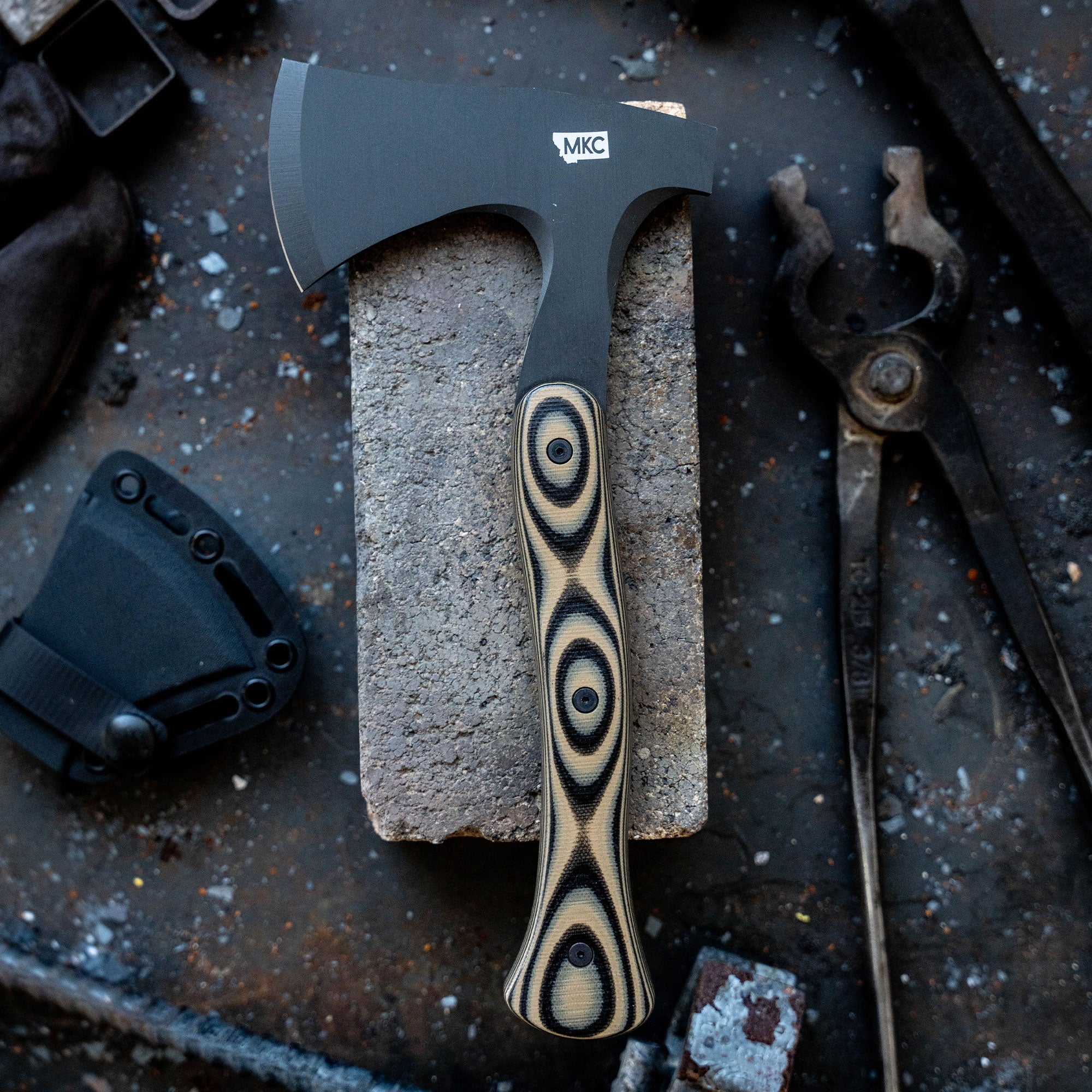 Alt text: "AEV Hellgate Hatchet from Secret Drop featuring a sleek, black blade and a striking striped handle, prominently displayed on a concrete block surrounded by workshop tools."