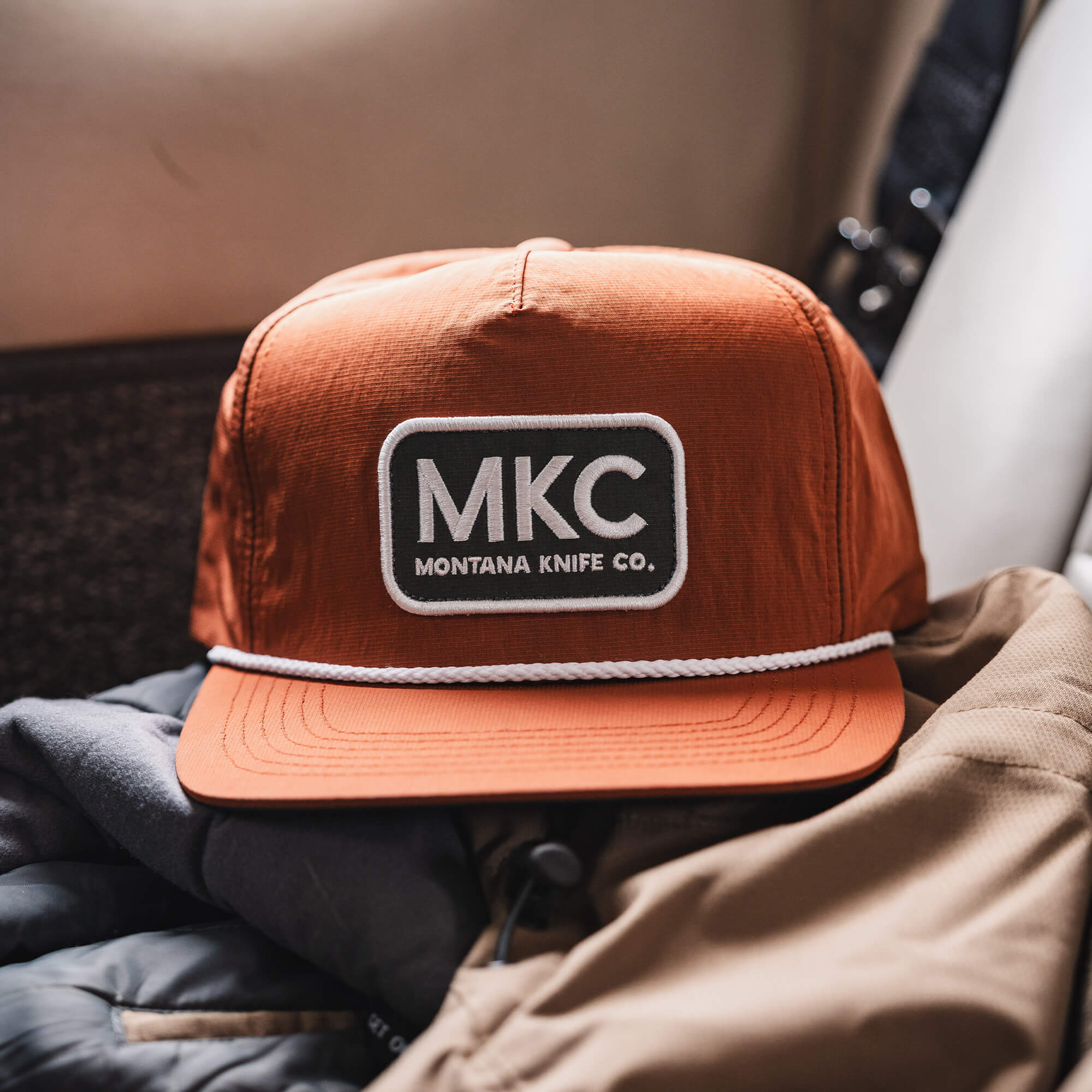 Rust-colored MKC River Rope Hat featuring a prominent Montana Knife Co. logo, resting on a jacket in a vehicle.