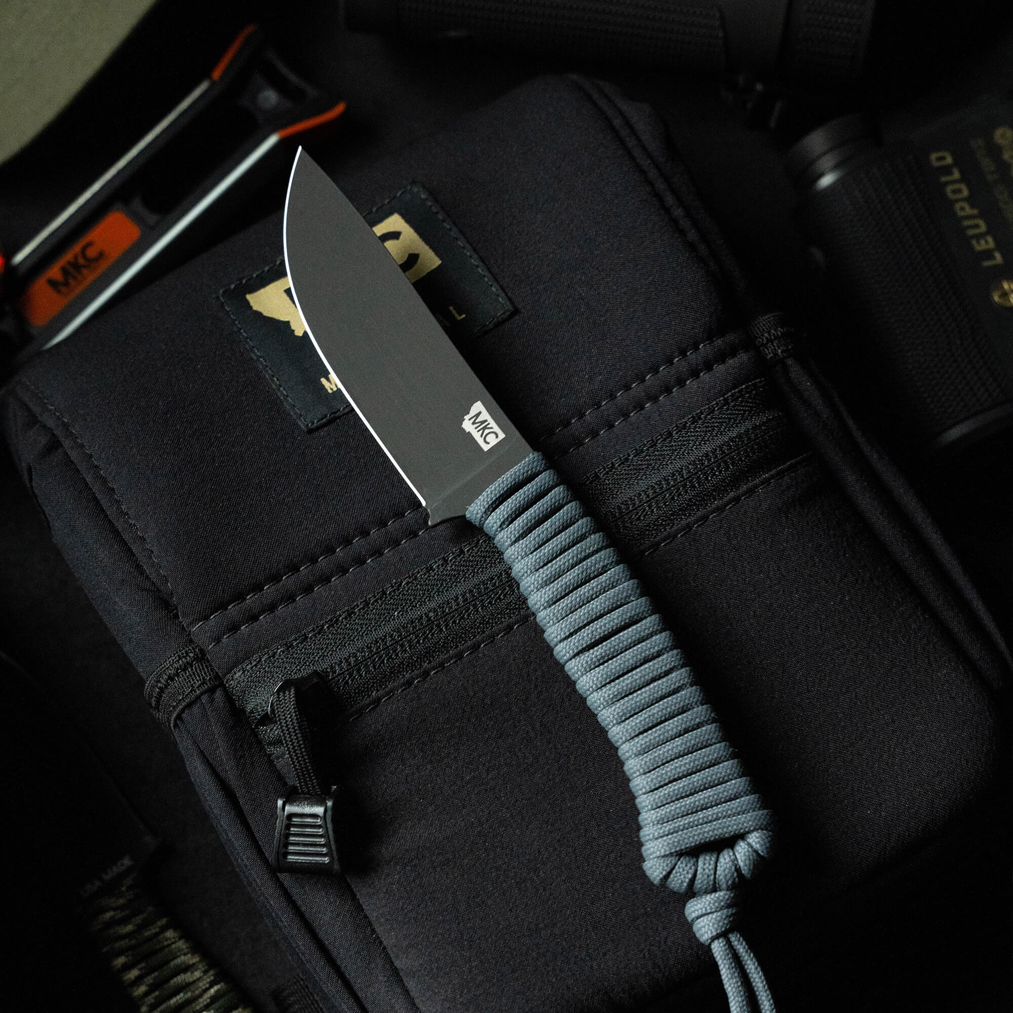Fixed blade knife with gray paracord wrapped handle placed on a tactical bag, surrounded by outdoor gear including a flashlight and another knife. The knife features a robust matte-black blade marked with the MKC logo.