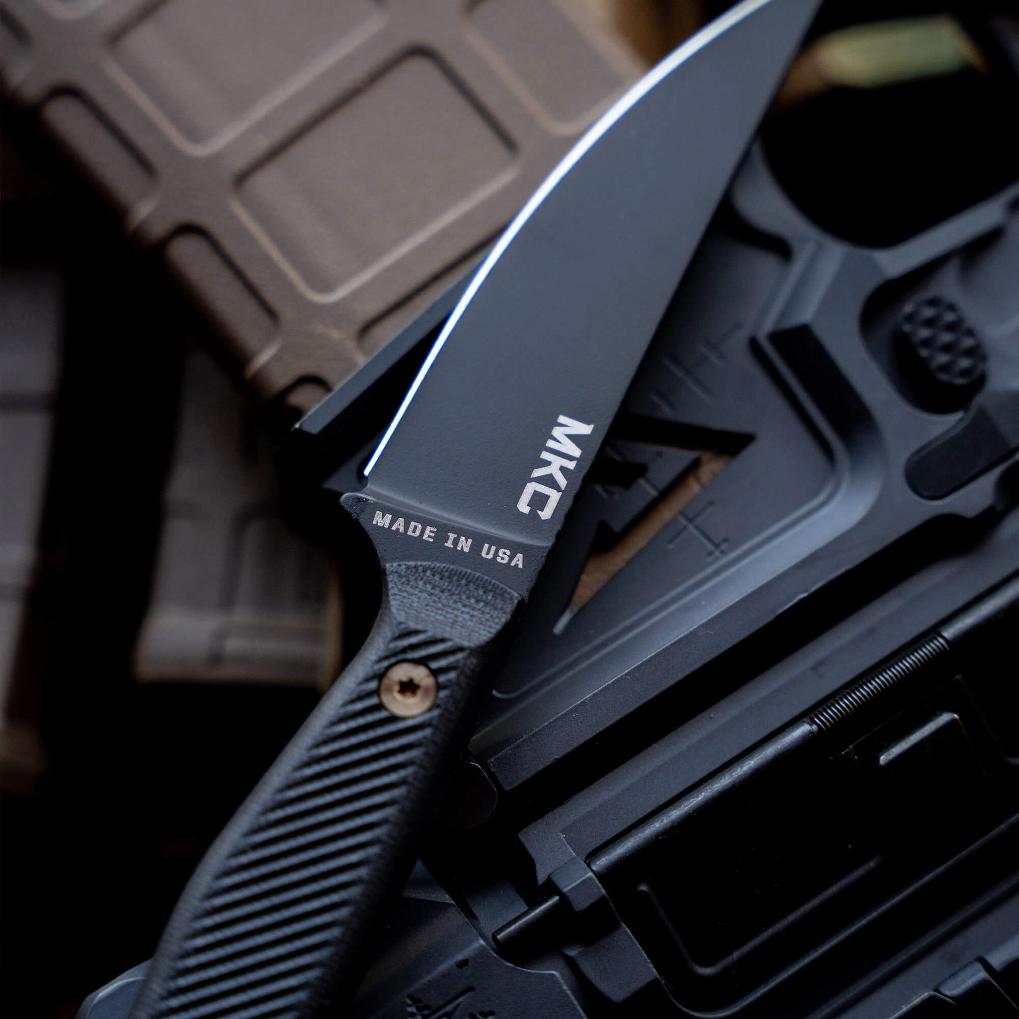 Close-up image of a black tactical knife with a blue line along the blade, labeled "MKC" and "MADE IN USA," resting on a textured assault rifle and ammo magazine.