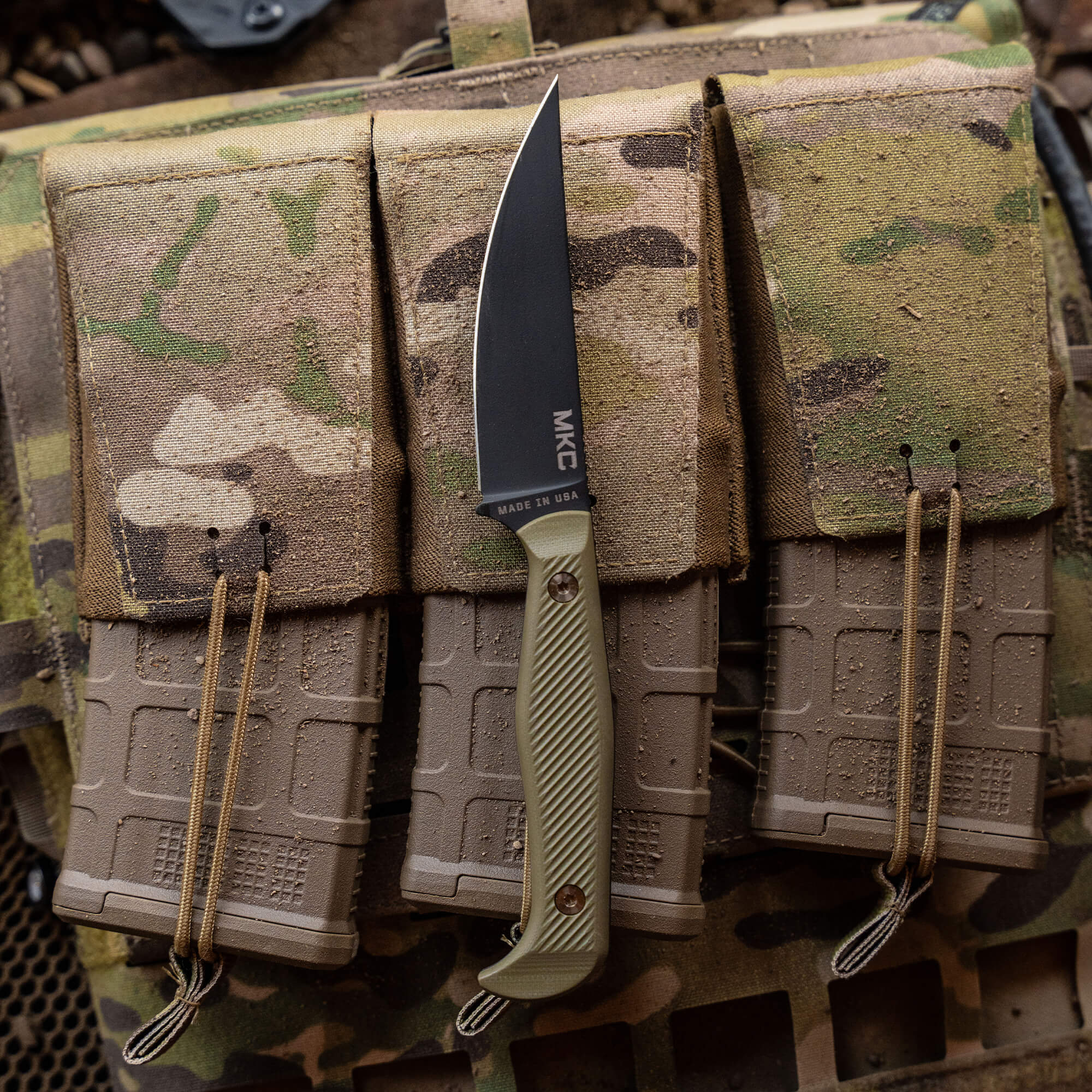 A close-up image of the BATTLE GOAT tactical knife in black and olive drab color, featuring a sleek, black blade and textured handle. The knife is nestled in a camouflage tactical vest with multiple pouches.
