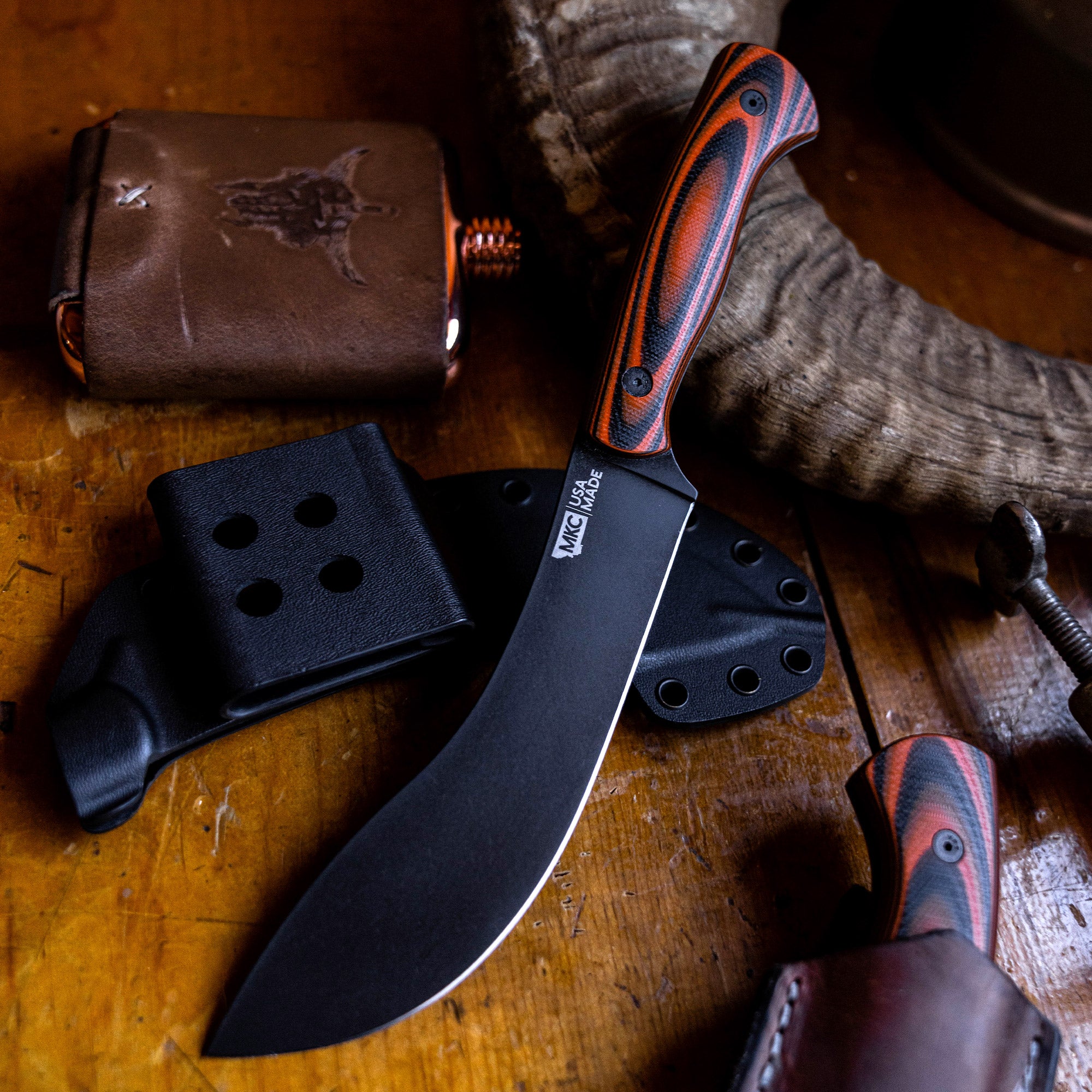 Alt text: "A close-up of the Beartooth Pro Skinning Blade with orange and black handle, displayed on a wooden surface alongside its black sheath and a vintage leather wallet. The curved, matte black blade is prominently featured, highlighting its