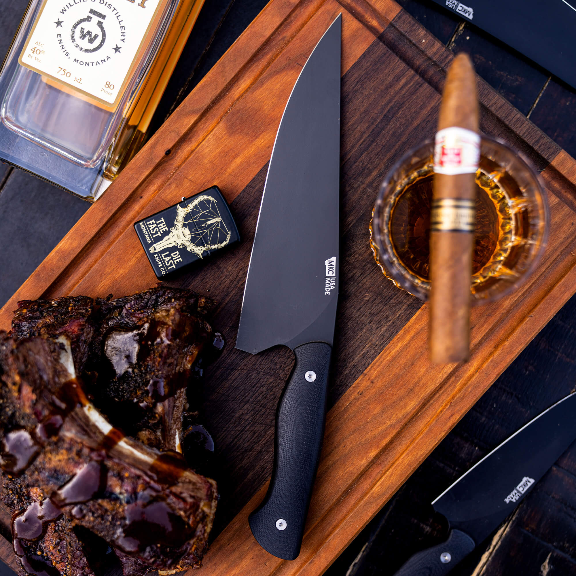 Alt text: "Elegant display of a Bighorn Chef black kitchen knife on a wooden board, accompanied by a cooked rib, a cigar in a glass ashtray, and a bottle of whiskey, evoking a luxurious culinary atmosphere."