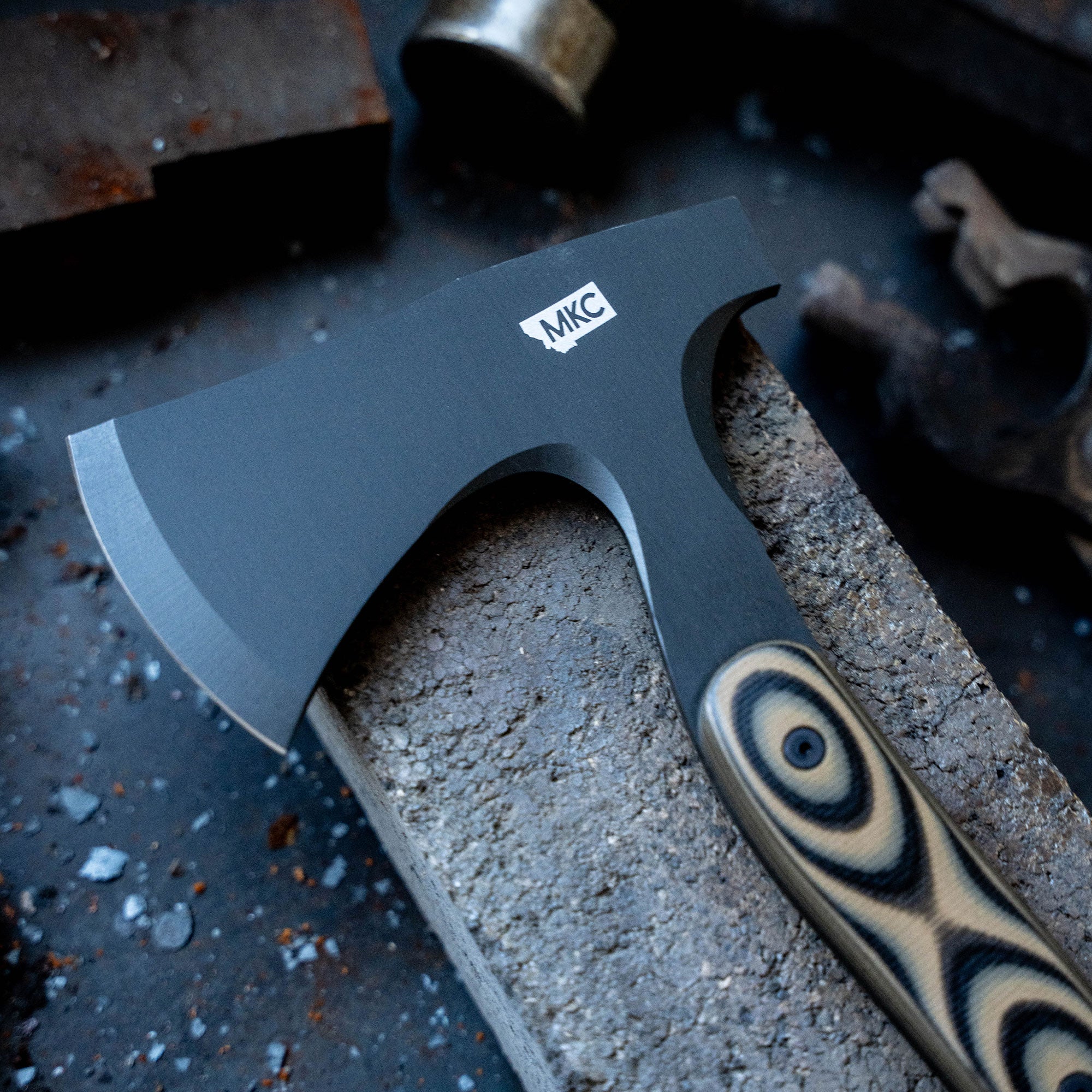 AEV Hellgate hatchet with a textured black blade and striped handle, displayed on a gritty, industrial metal surface.