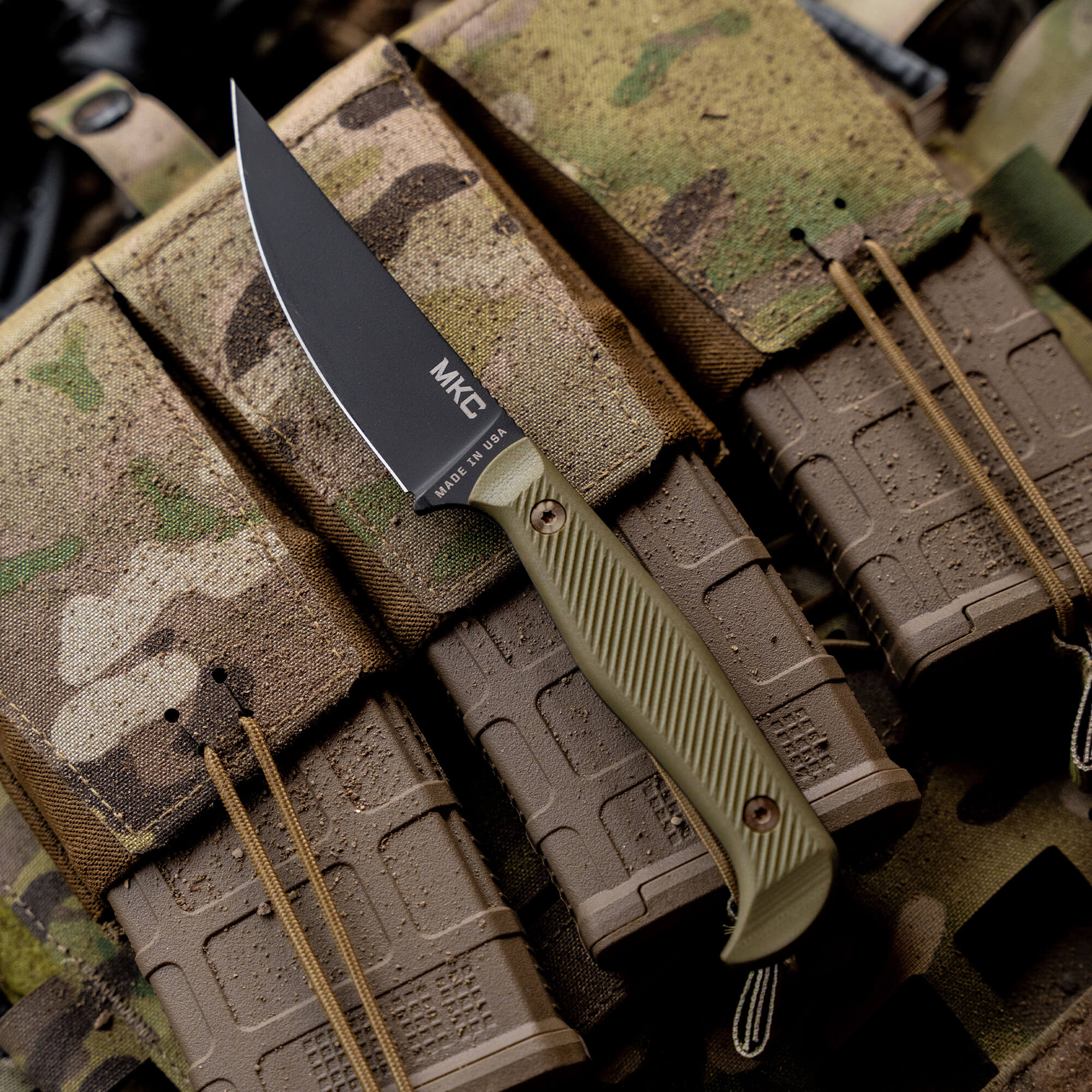 Alt text: "Close-up of a BATTLE GOAT BLK/OD tactical knife by MKT, featuring a black blade and olive drab handle, displayed on a camouflaged tactical gear background."