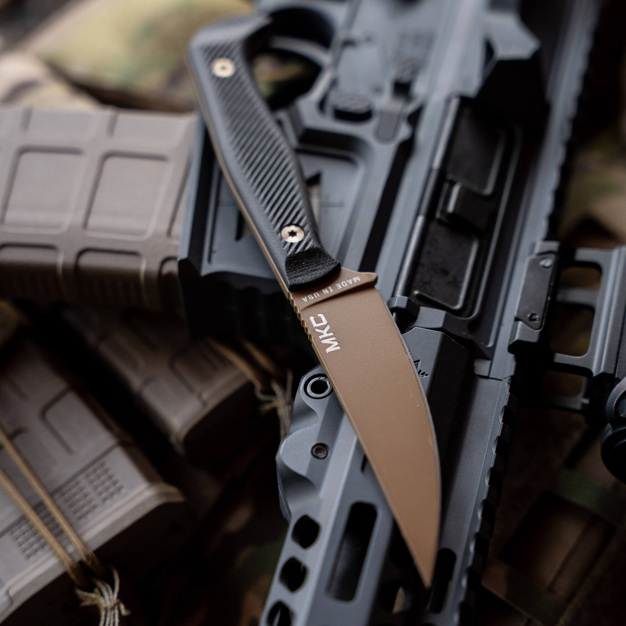 Alt text: "Close-up of a Coyote Black (COY/BLK) Battle Goat tactical knife with a tan blade and black handle, resting on a rifle and tactical gear."
