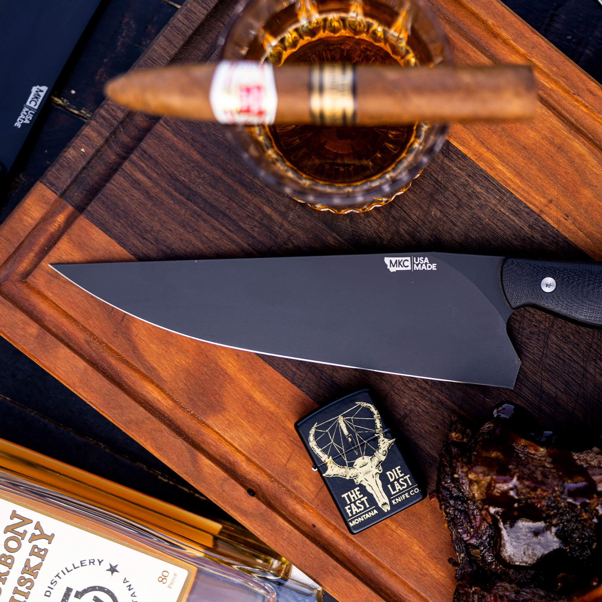 Alt text: A sleek Bighorn Chef black kitchen knife displayed on a wooden board, surrounded by upscale accessories including a lit cigar resting in a glass of bourbon, a stylish lighter, and elegant packaging from a bourbon distillery.