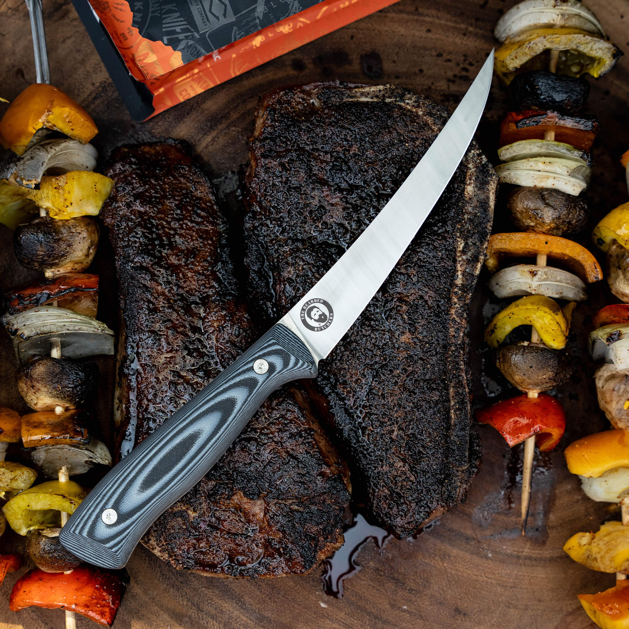 Alt text: "Bearded Butchers boning butcher knife with a black and gray handle, displayed on a wooden board alongside a cut of well-cooked beef brisket and colorful grilled vegetables including mushrooms, peppers, and onions."