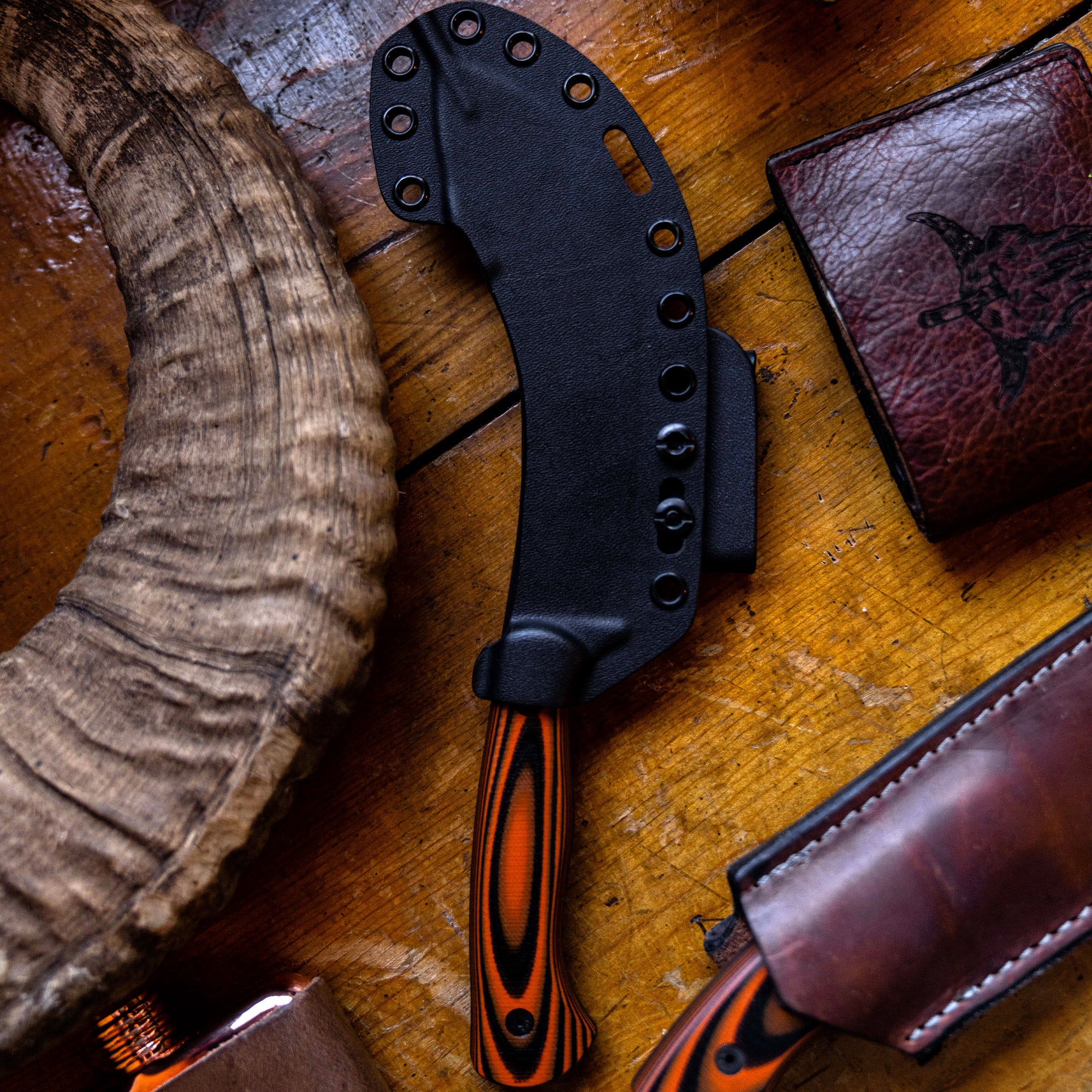 Alt text: "Close-up view of Beartooth Pro Skinning Blade with orange and black handle, displayed on a wooden surface next to a leather wallet and antler piece, comeplete with a black sheath."