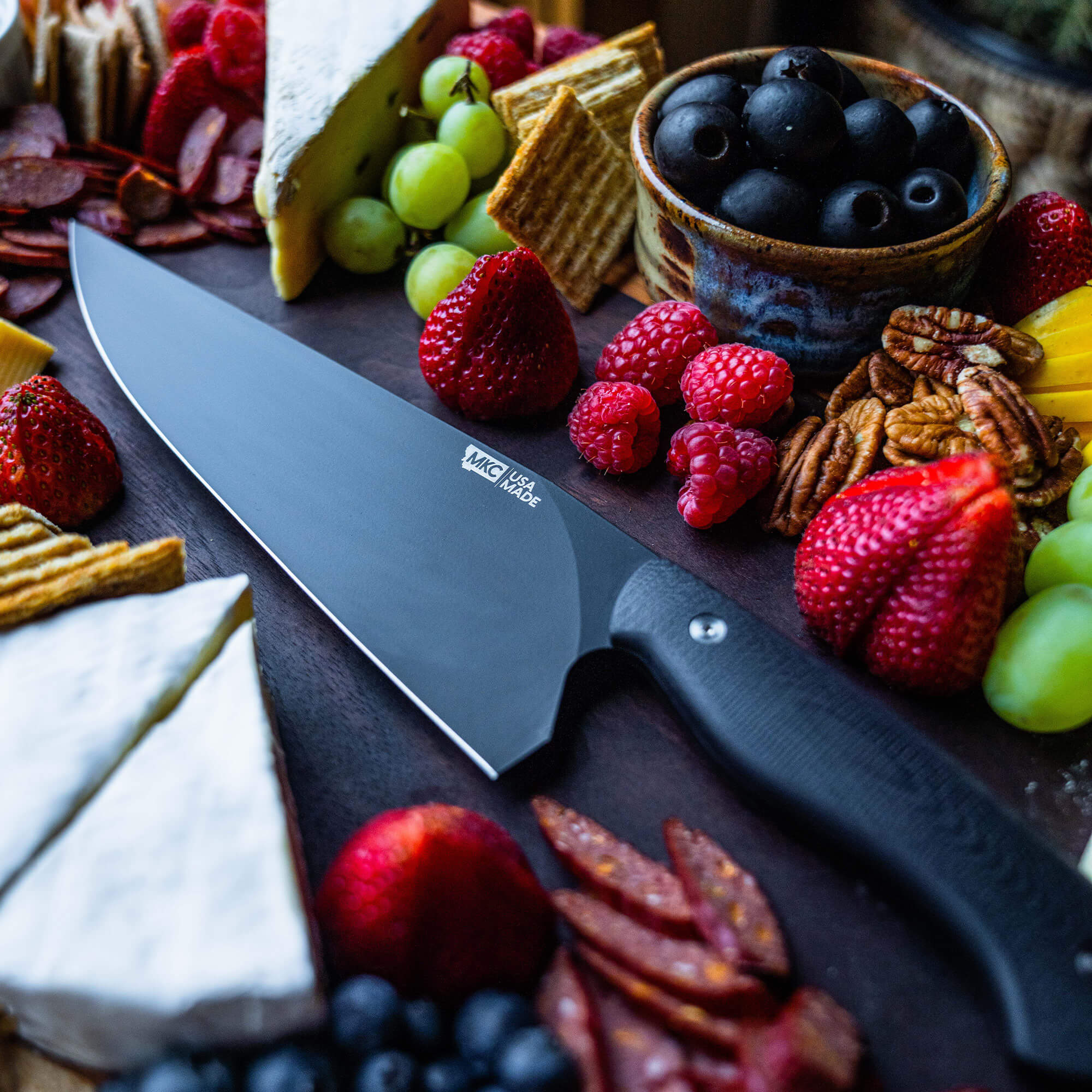 Alt text: BIGHORN Chef - Black professional chef's knife displayed on a dark wood board surrounded by an assortment of gourmet cheeses, fruits, nuts, and charcuterie, highlighting its sleek blade and ergonomic black handle.