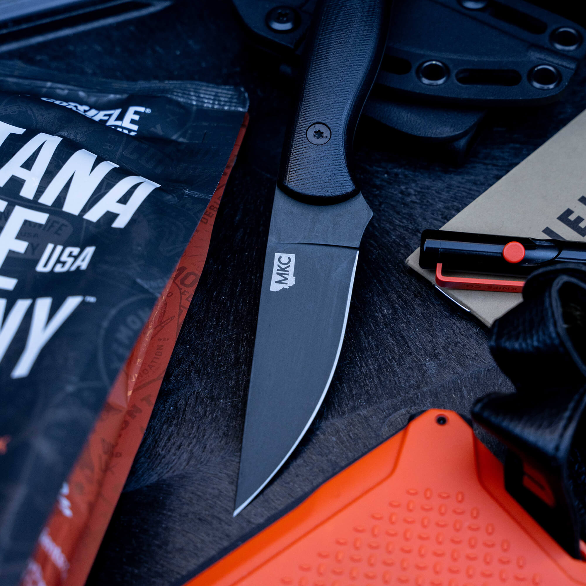 Alt text: "Close-up view of the BLACKFOOT 2.0 tactical knife with black handle and silver blade, displayed on a dark textured surface surrounded by survival gear including an orange emergency whistle, a sheath, and a map."